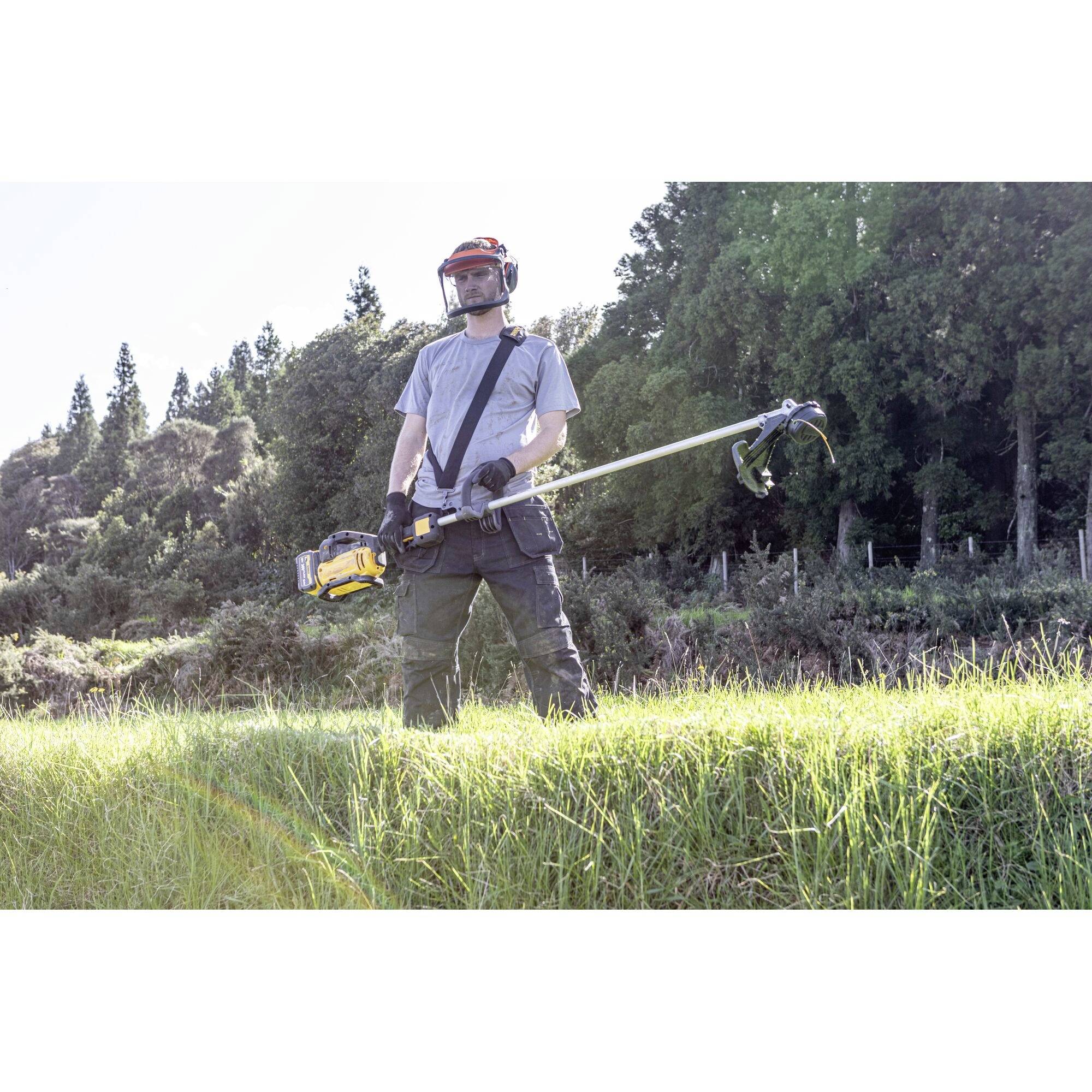 A person in protective clothing is mowing grass with a strimmer on a meadow in front of trees.