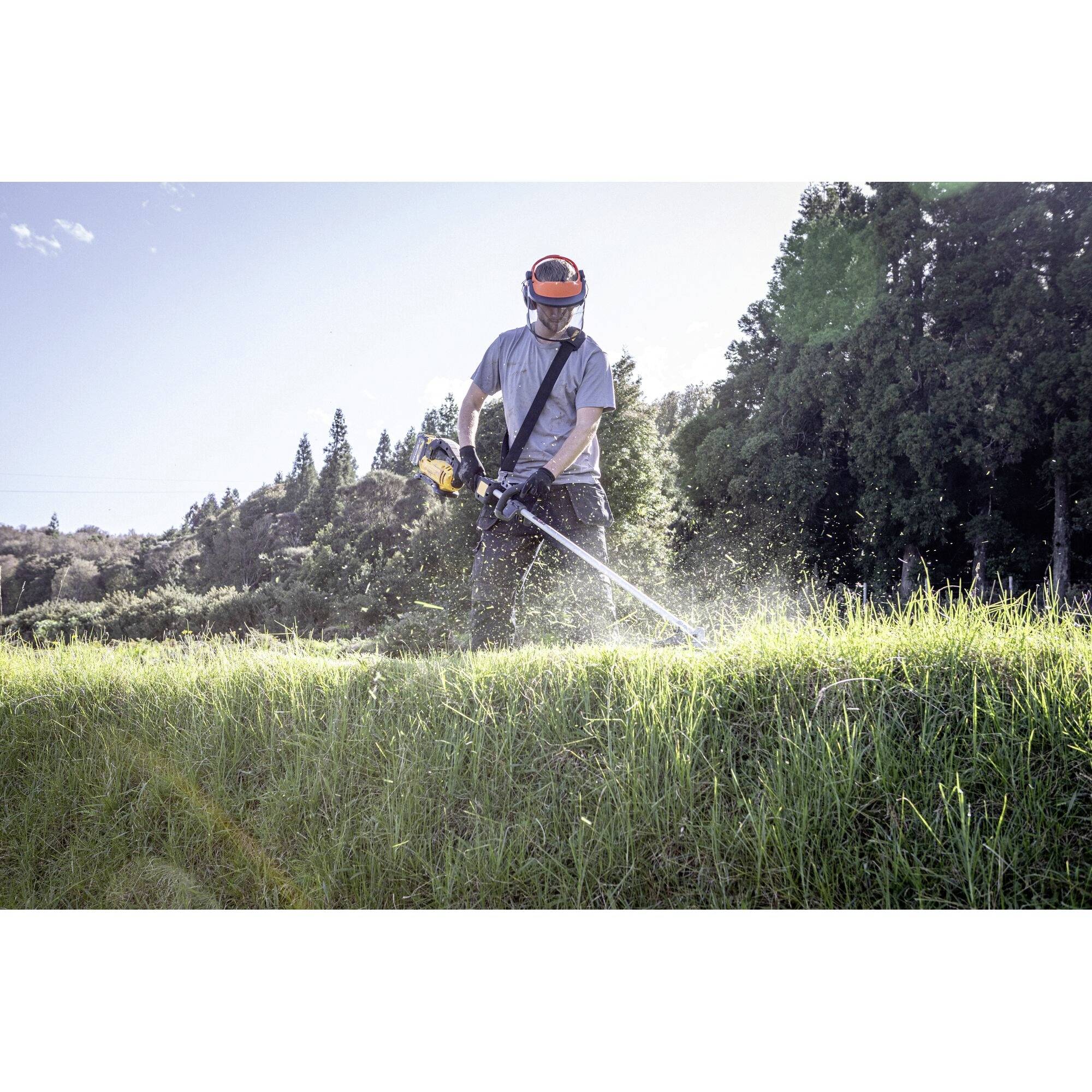 A person wearing a protective helmet is strimming grass on a meadow with a motorised brush cutter. Background: Trees and blue sky.
