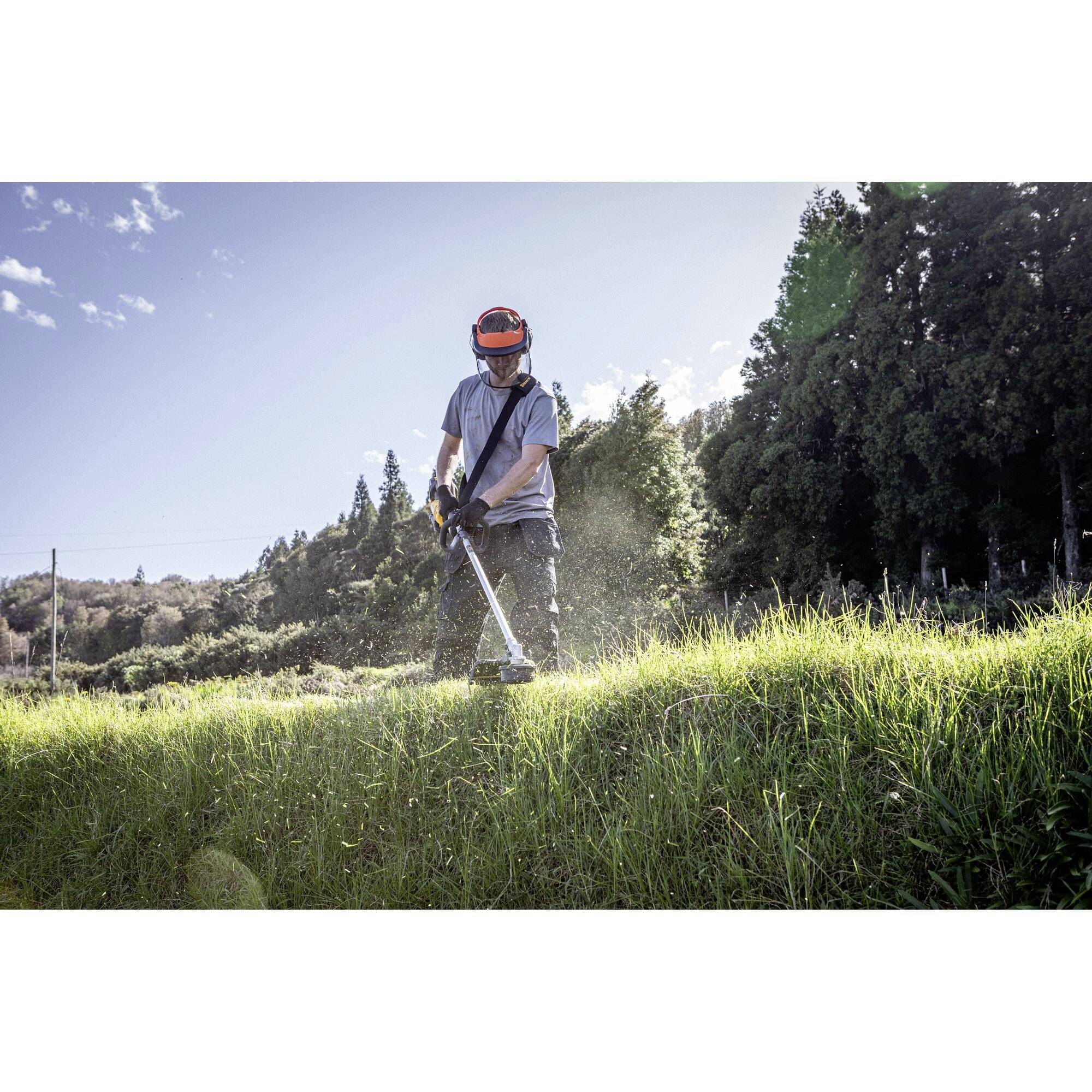 A person is mowing grass with a petrol strimmer. Trees and a clear sky can be seen in the background.
