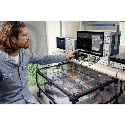 A man operates an electronic measuring station, examining circuits in a Perspex enclosure, with oscilloscopes in the background.