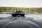 A black remote-controlled boat with an antenna is sailing on a calm lake. A forest is visible in the background.