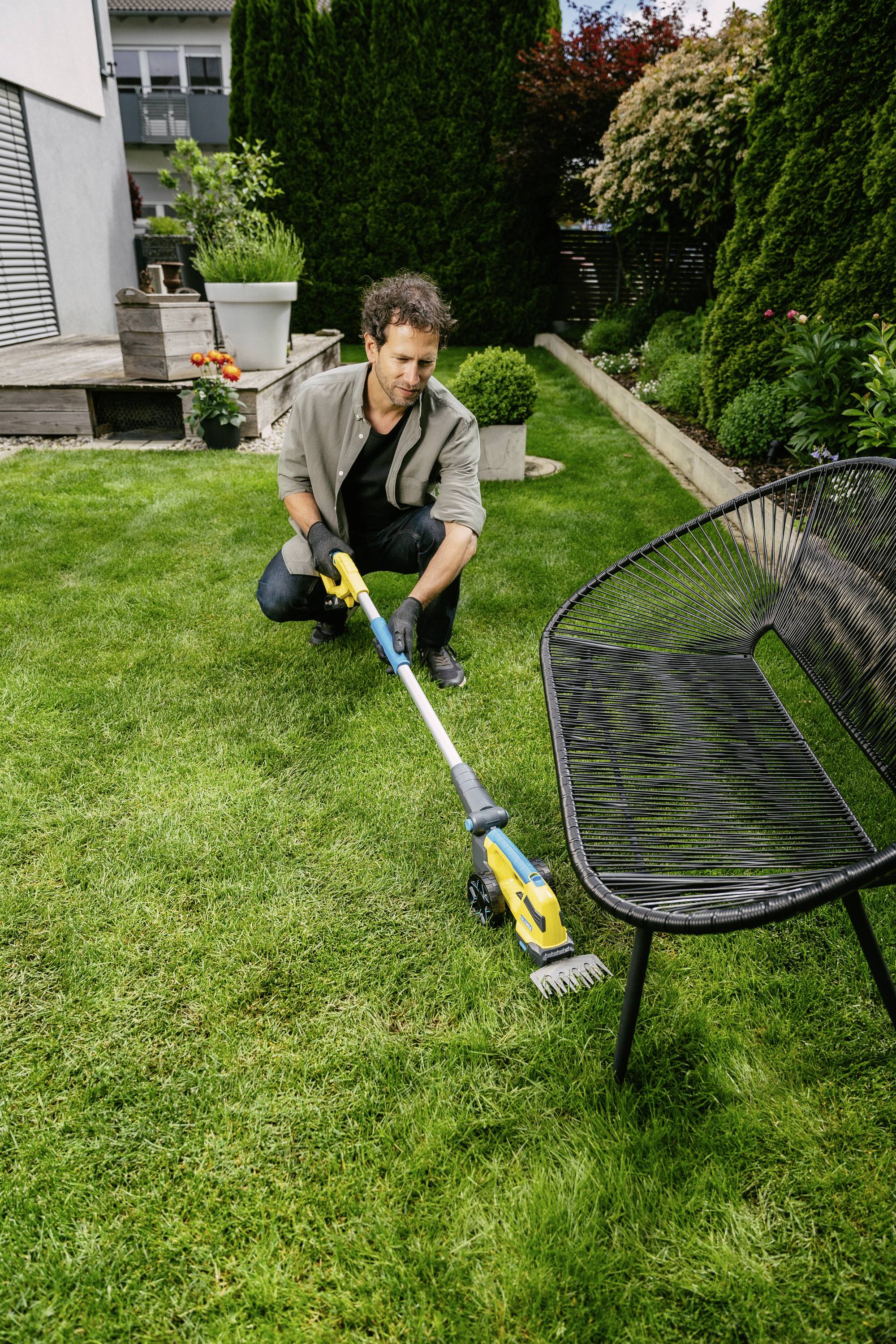 A man is using an electric garden tool to maintain the lawn next to a bench. Plants and a house are visible in the background.
