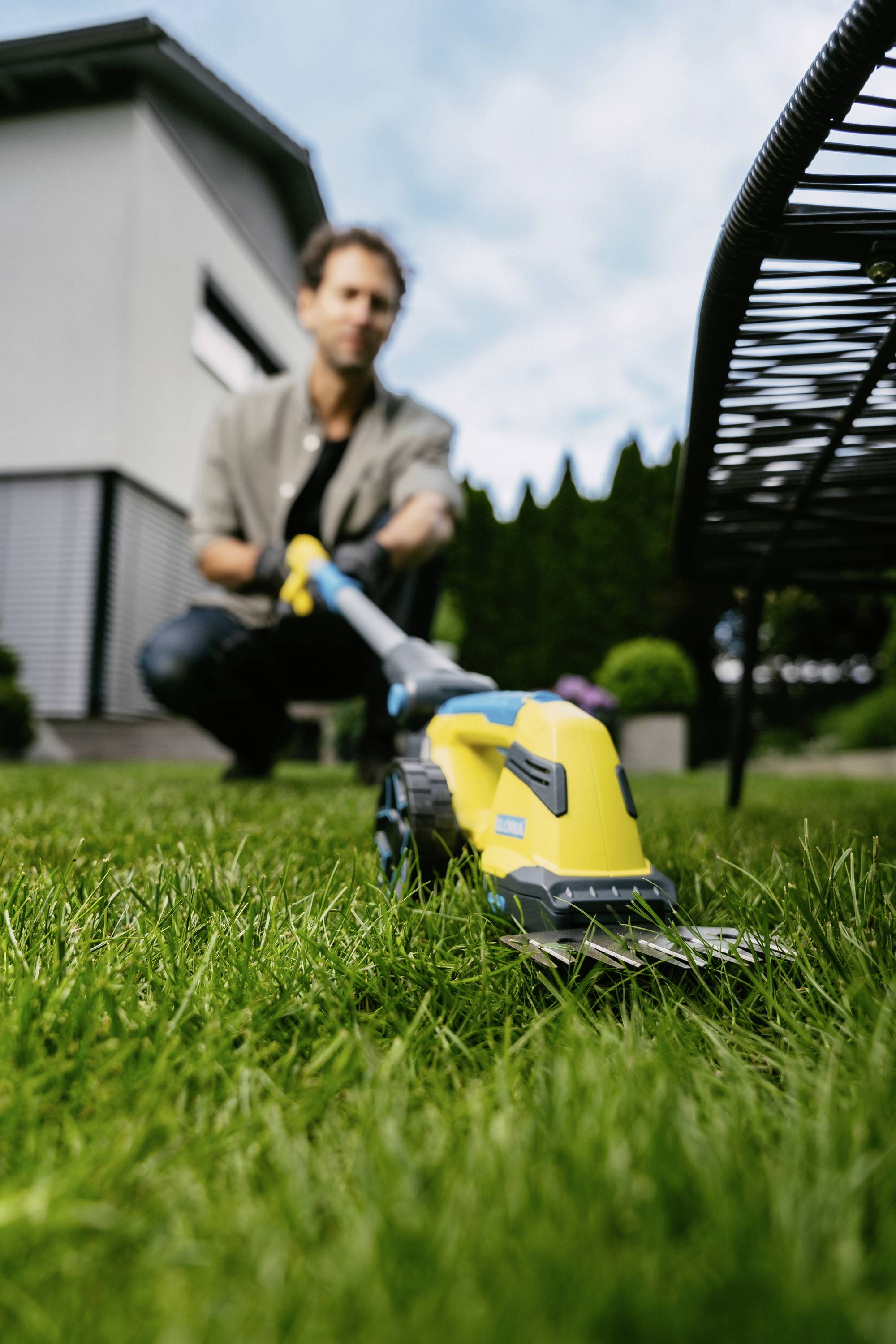A person is using a yellow and blue lawn trimmer in the garden. A modern house can be seen in the background.