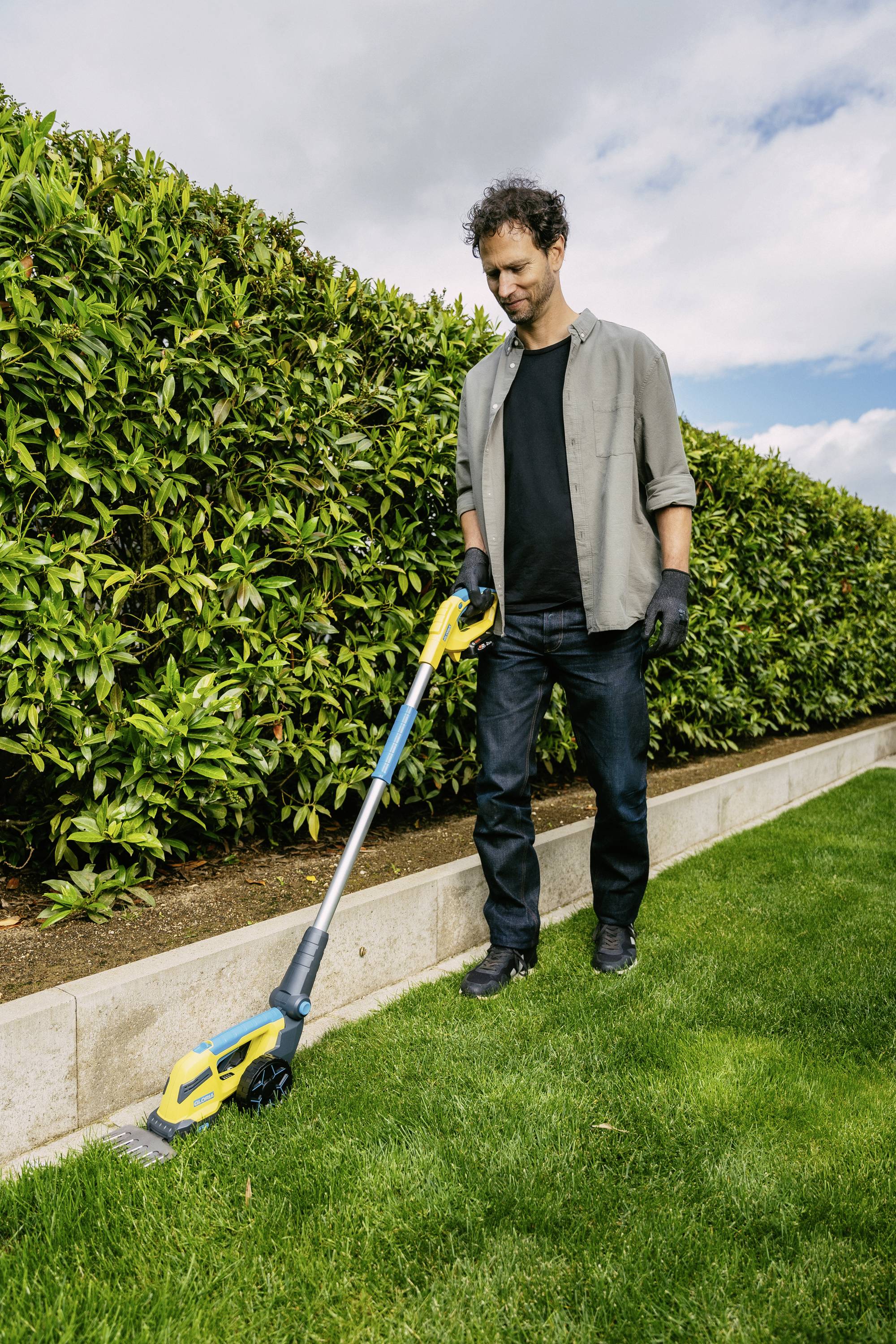 A man is trimming the lawn edge along a hedge with an electric hedge trimmer. He is wearing gloves and looking concentrated.