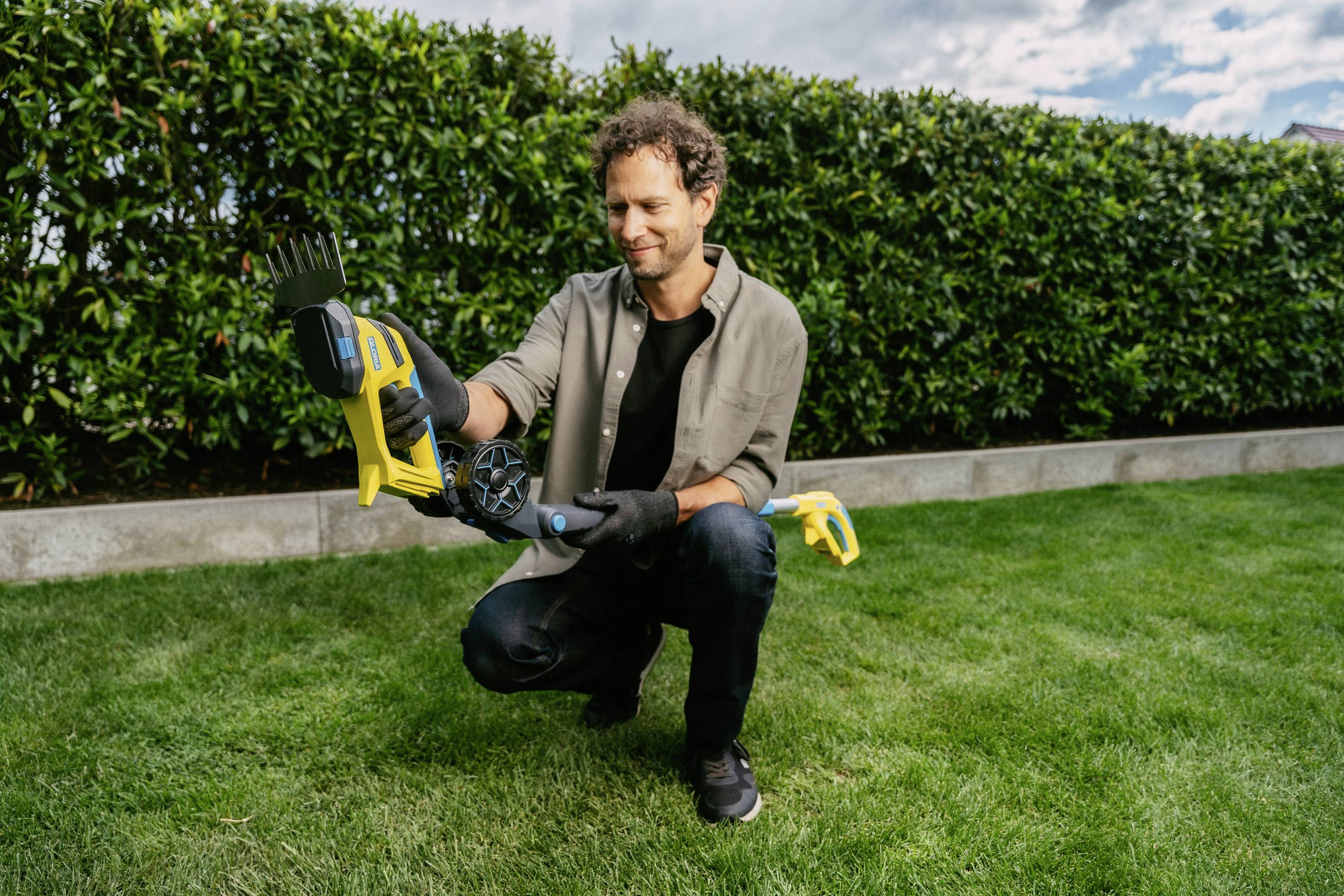 A man is kneeling on a green lawn and holding an electric hedge trimmer. A hedge is visible in the background.