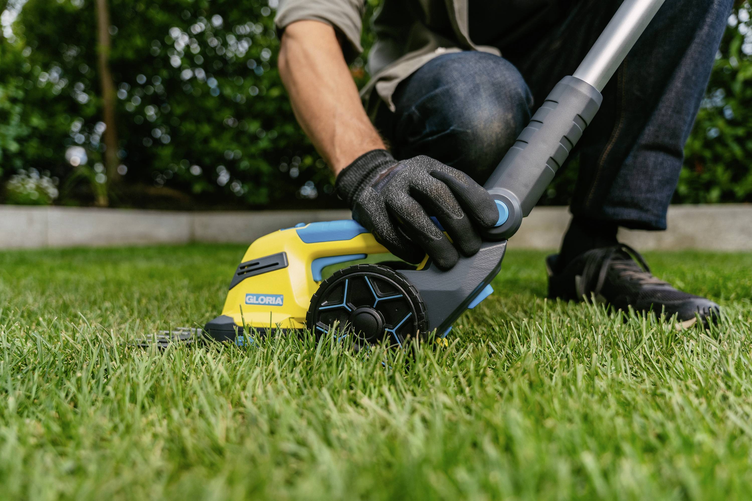 A person is kneeling on the lawn and trimming grass with a yellow lawn strimmer.