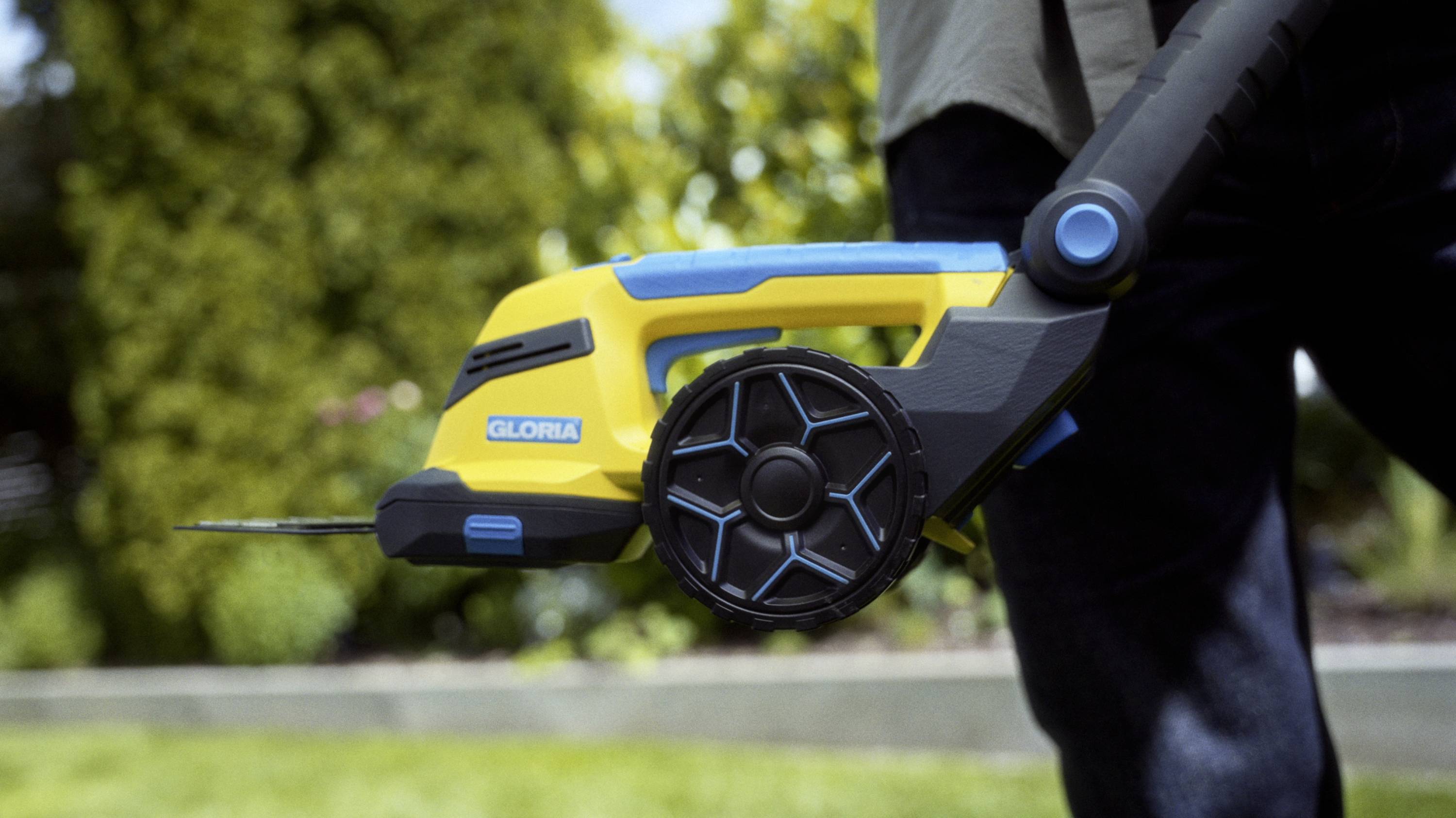 A person is holding a yellow electric lawn trimmer with a black handle. A garden is visible in the background.
