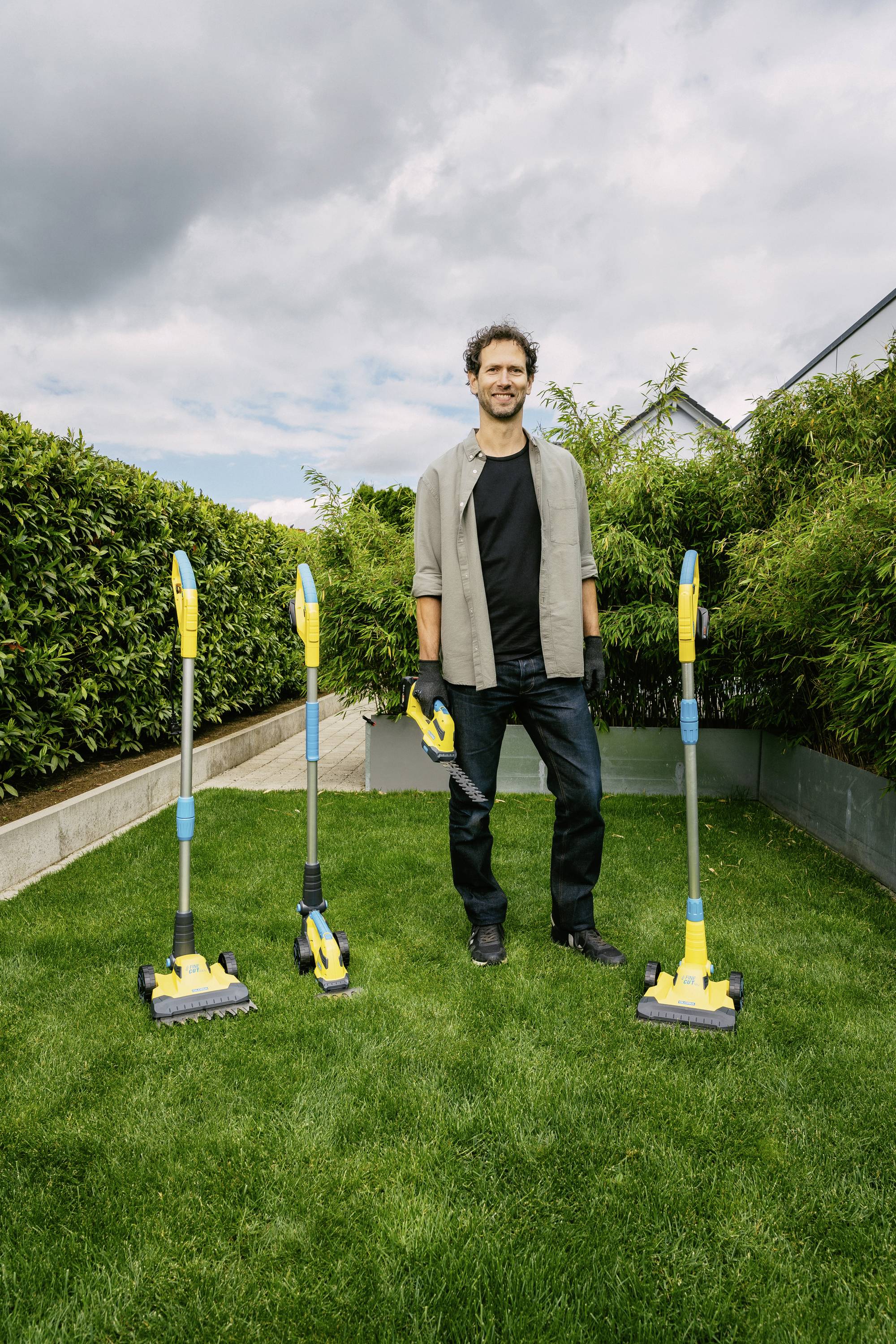 A person is standing smiling on green grass in a garden, surrounded by three yellow and blue cleaning tools, with hedges in the background.