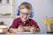 A smiling boy with glasses and blue headphones is playing on a smartphone at a table. Books and a pencil holder are lying beside him.