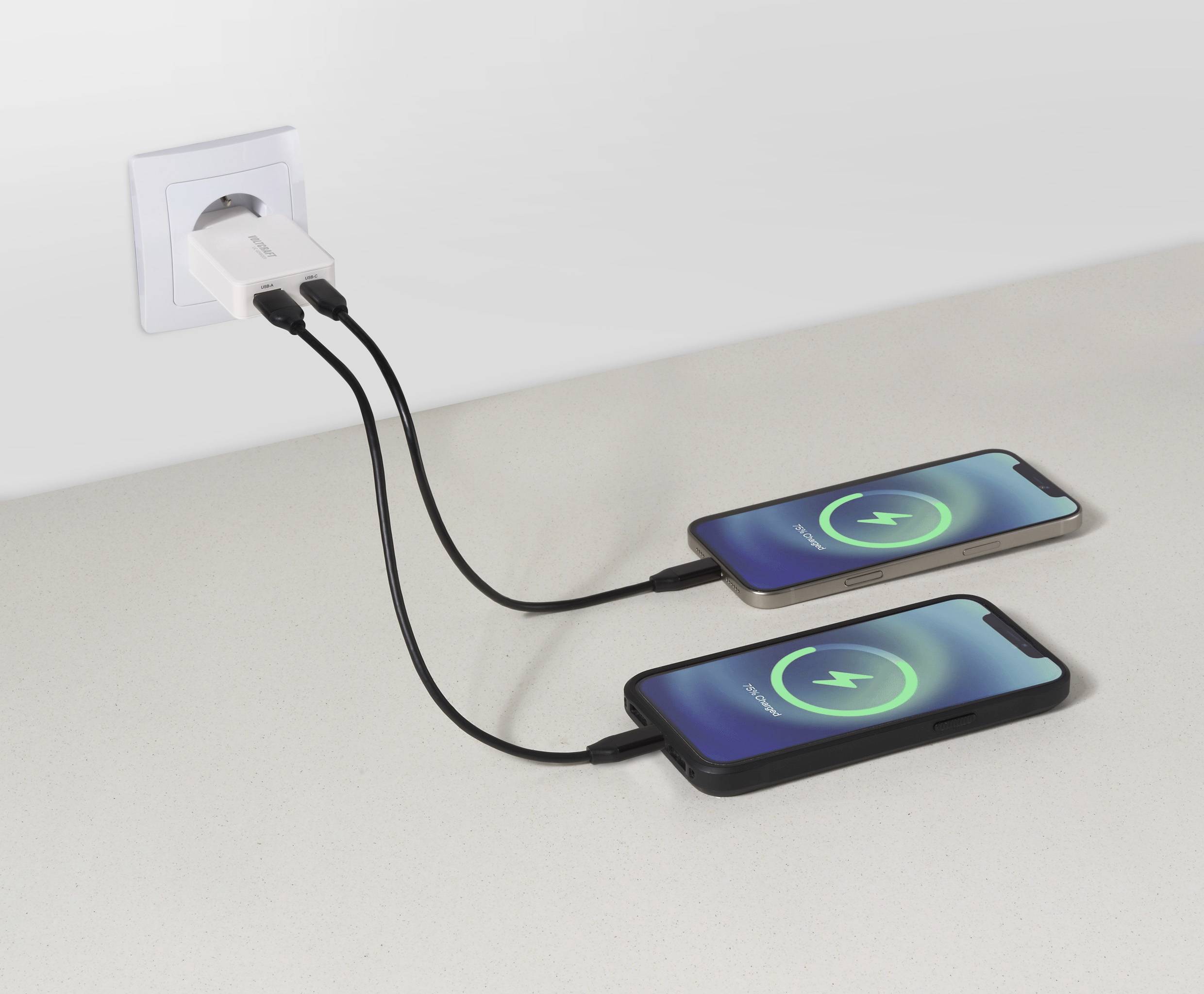 Two smartphones on a table connected to a wall adapter, both showing a green charging symbol on their screens, indicating active charging.