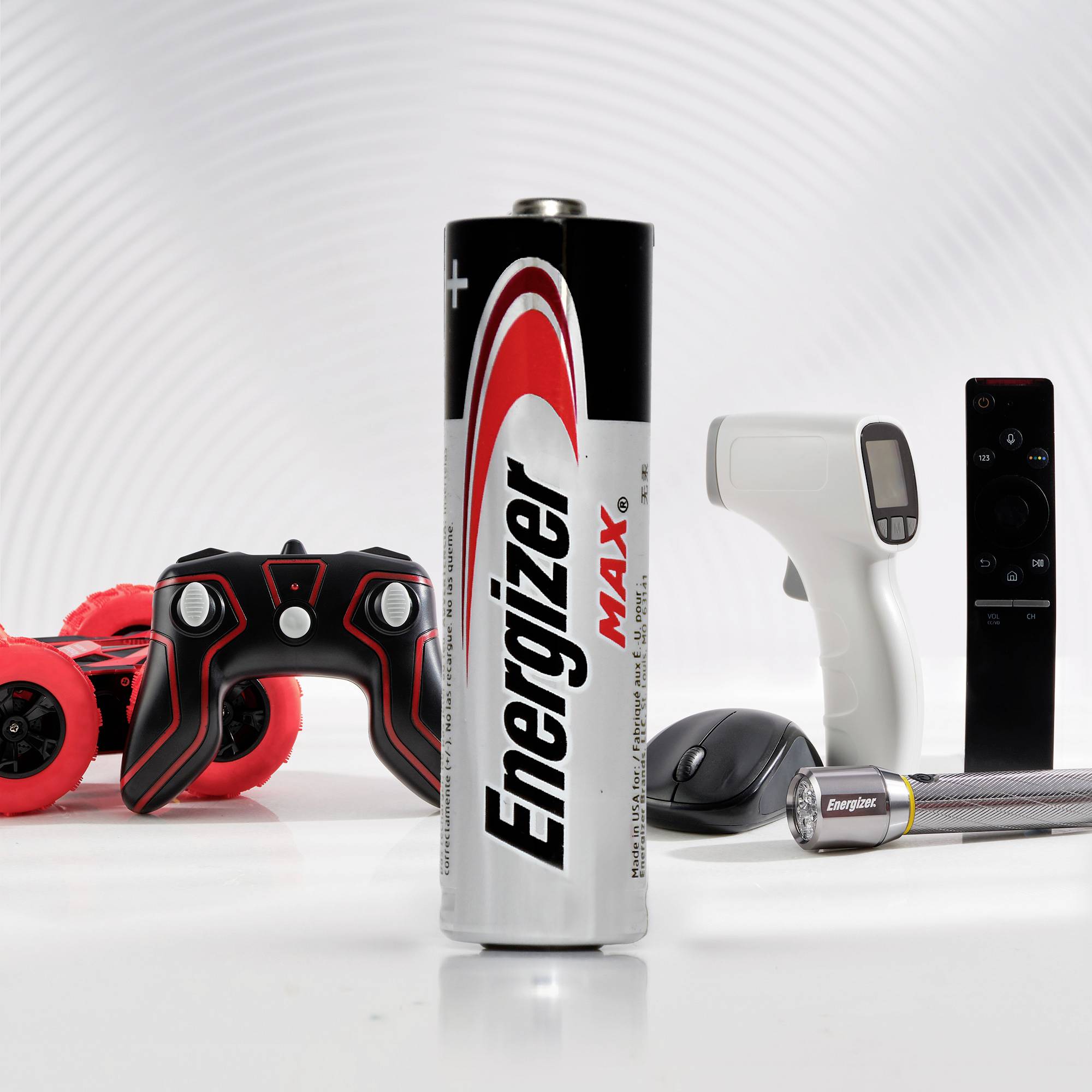 An upright AA battery is in the foreground. In the background, a toy controller, a thermometer, a remote control, another battery, and a toy car can be seen.