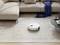 A white robotic vacuum cleaner is cleaning a beige carpet next to a beige sofa and a coffee table with a cup of coffee and a blue scarf.
