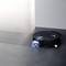 A black robot vacuum cleaner with copper-coloured accents is cleaning the floor in front of a light grey wall.