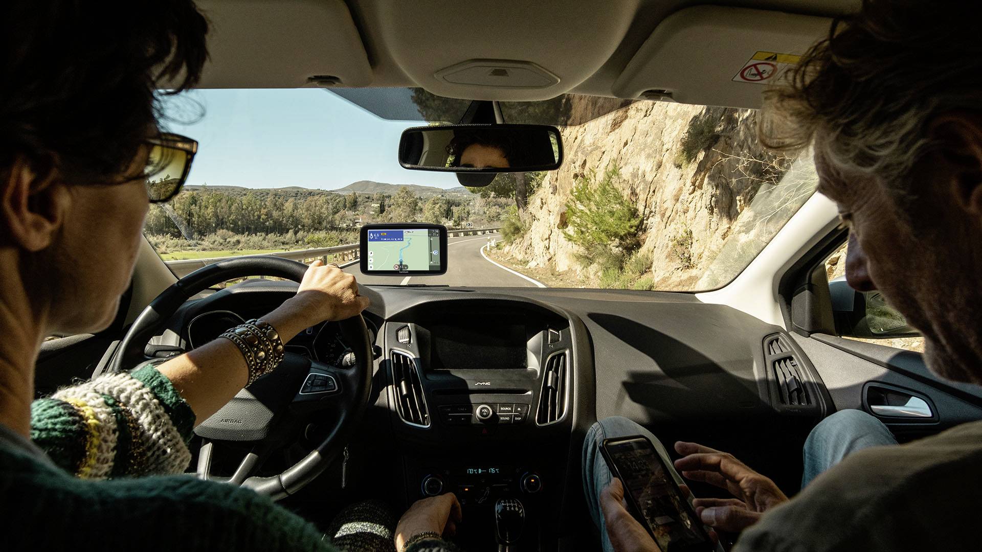 Two people are driving in a car on a winding country road. The driver is pointing at a satellite navigation device showing a route.