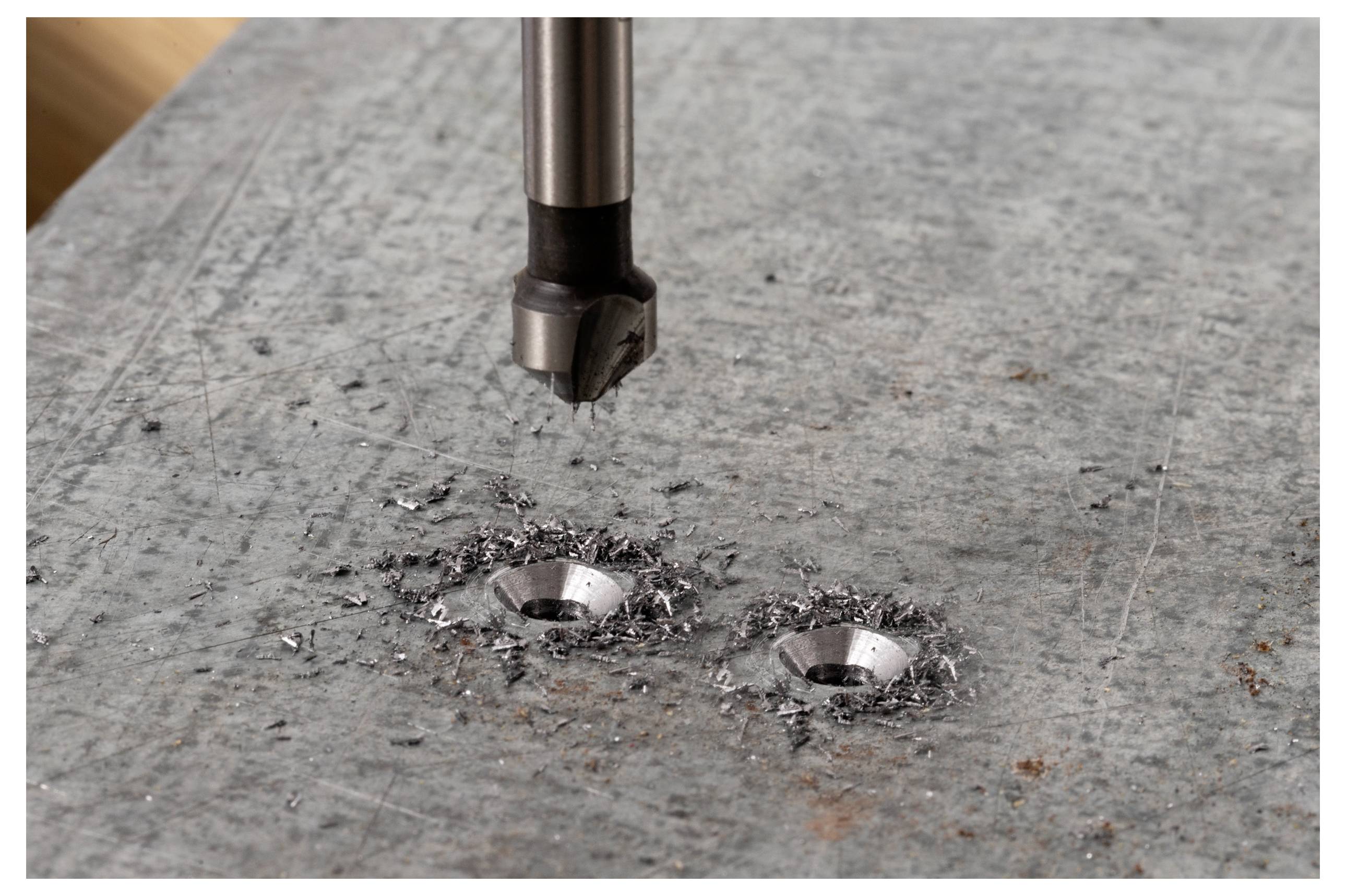 A drill bit is positioned above two freshly drilled holes in a metal surface, surrounded by metal shavings.