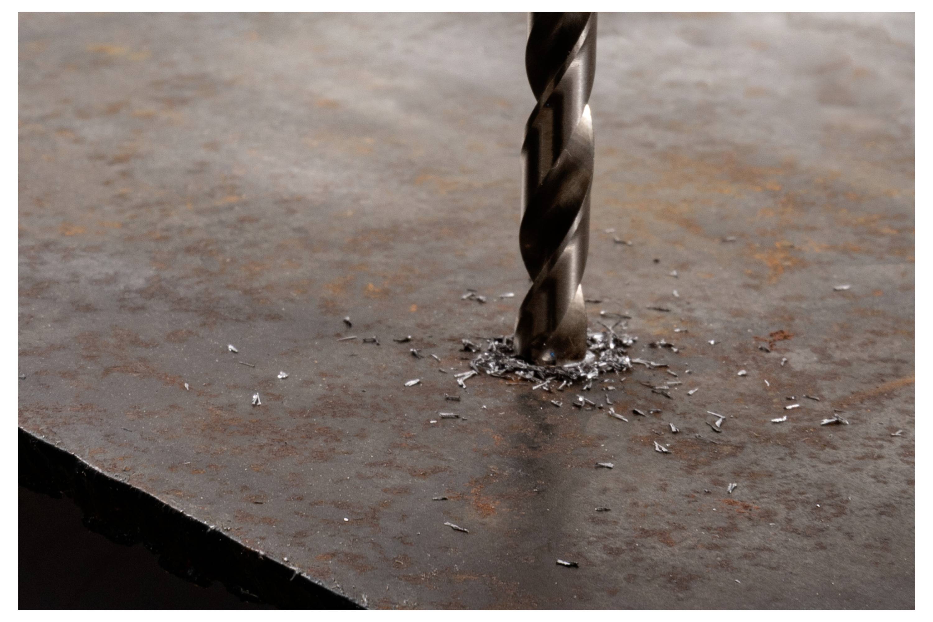 A metal drill bit actively drills through a rusted steel plate, producing small metal shavings around the hole, illustrating metalworking.