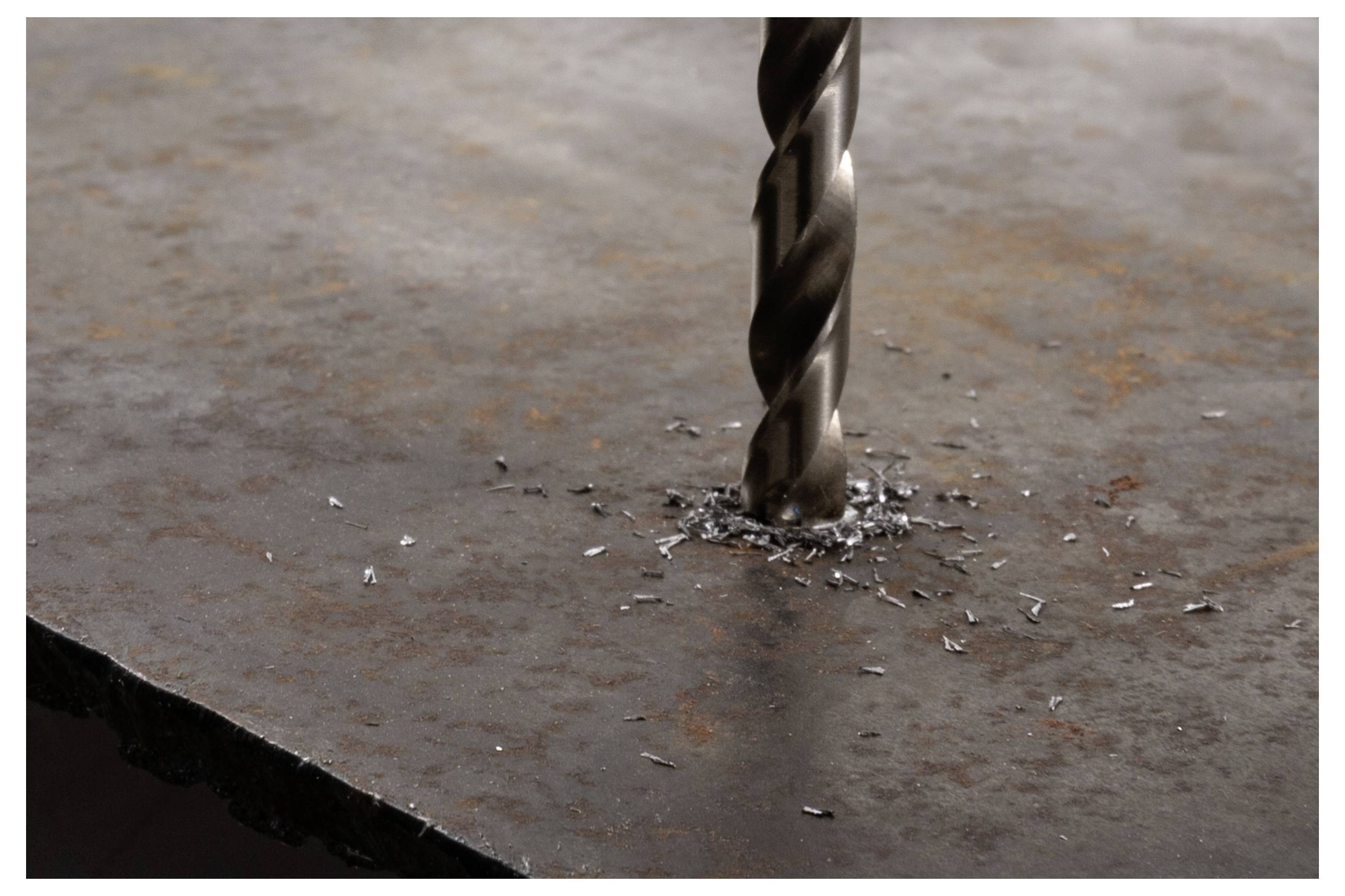 A metal drill bit is drilling through a rusted metal surface, creating spirals of metal shavings around the hole.