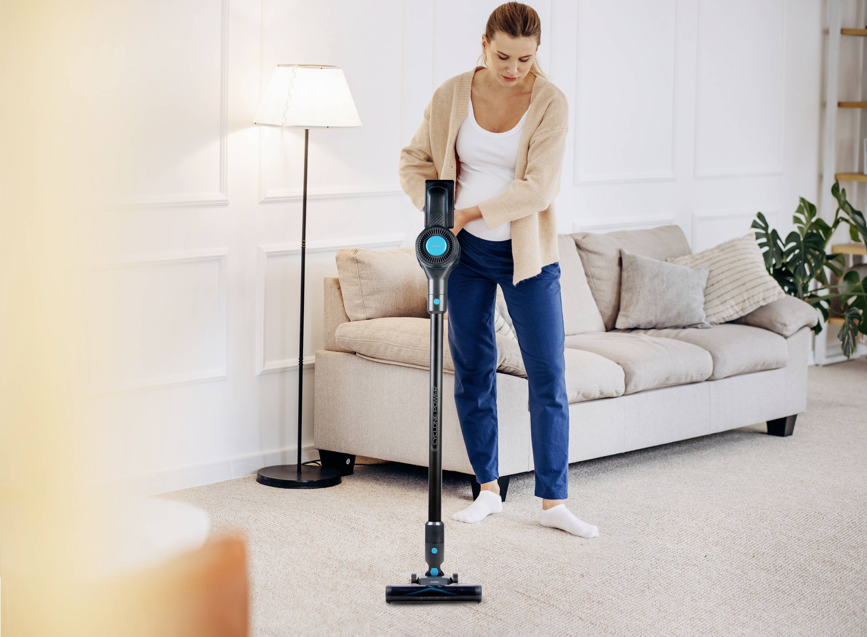 A person is using a cordless vacuum cleaner on a beige carpet in a cosy living room. A sofa and a lamp are visible in the background.