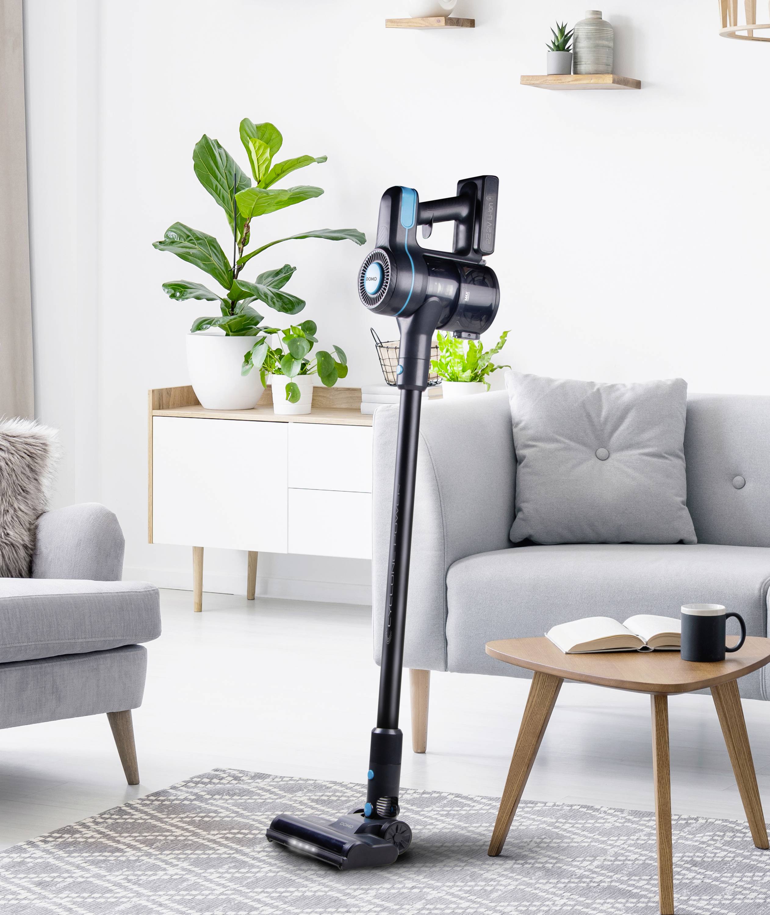 A cordless vacuum cleaner stands in a modernly furnished living room. In the background, an armchair, a table with a cup and book, and a houseplant can be seen.