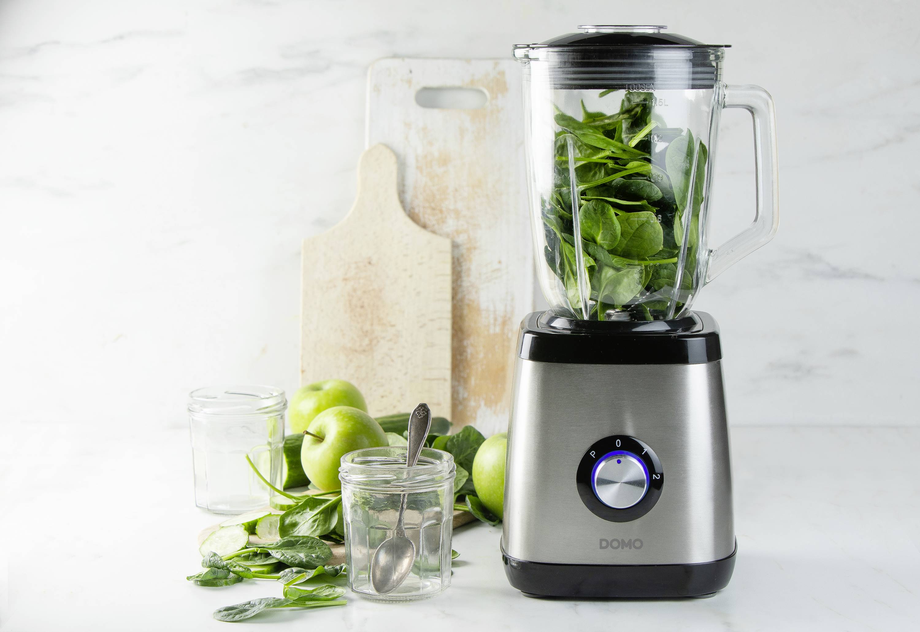 A blender with a glass container, filled with green leafy vegetables. In the background are apples, a wooden chopping board, and empty glasses.