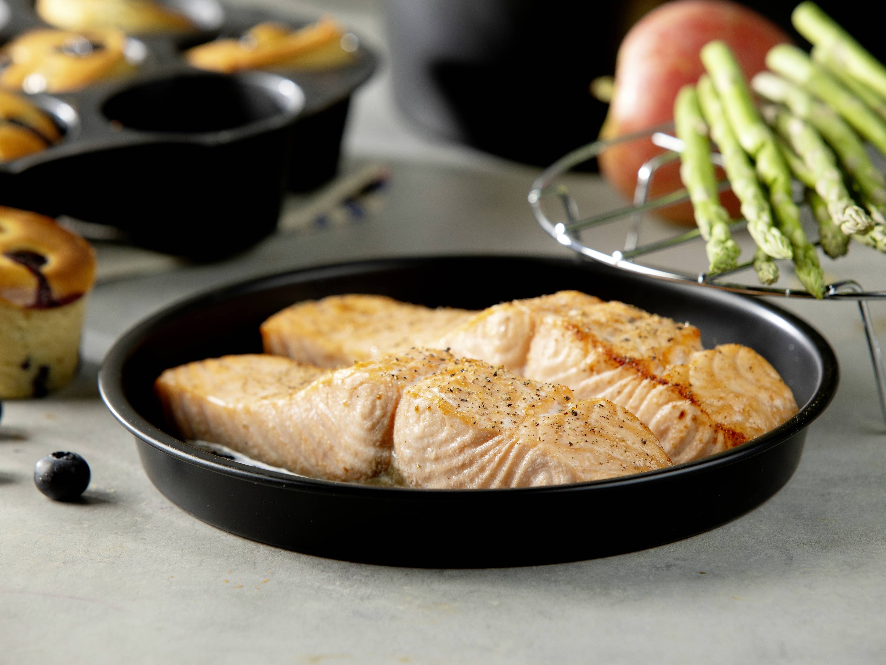 Baked salmon in a round pan, garnished with pepper. Asparagus, muffins, and an apple visible in the background.
