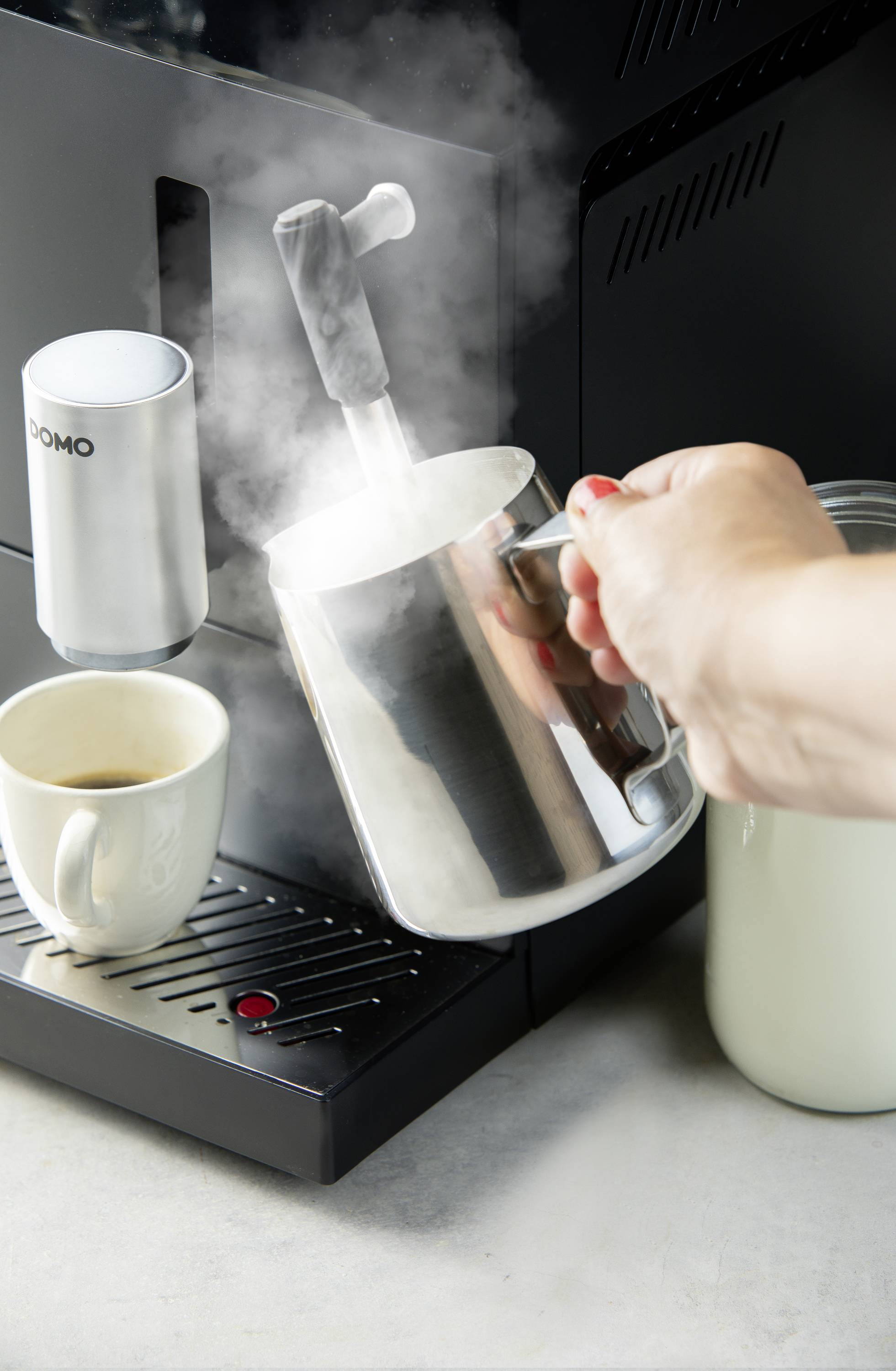 A person is frothing milk using the steam wand of a coffee machine. A white cup is positioned beside it.