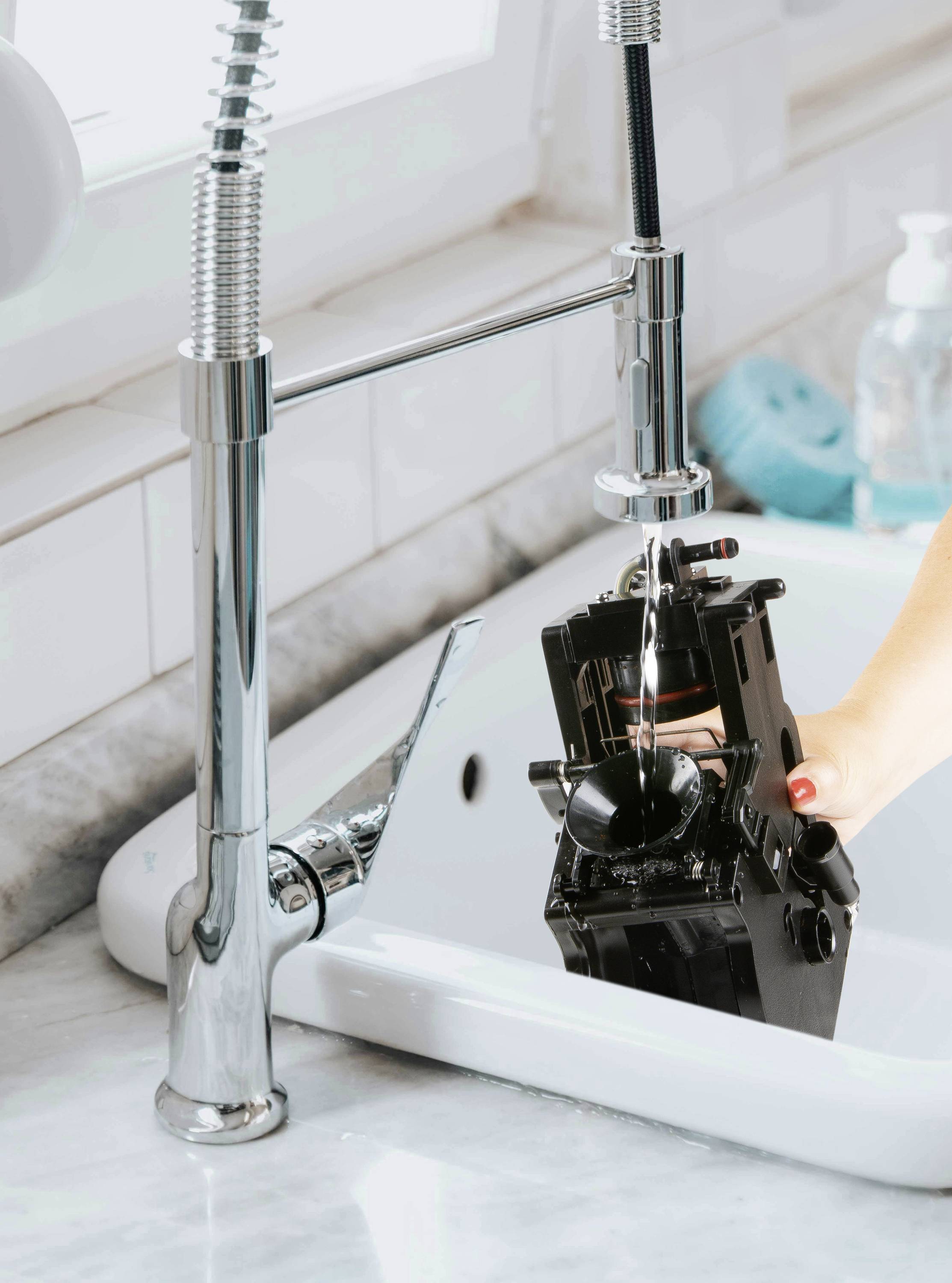 A person is cleaning a black camera under running water in a sink. Tiles and a soap dispenser bottle are visible in the background.