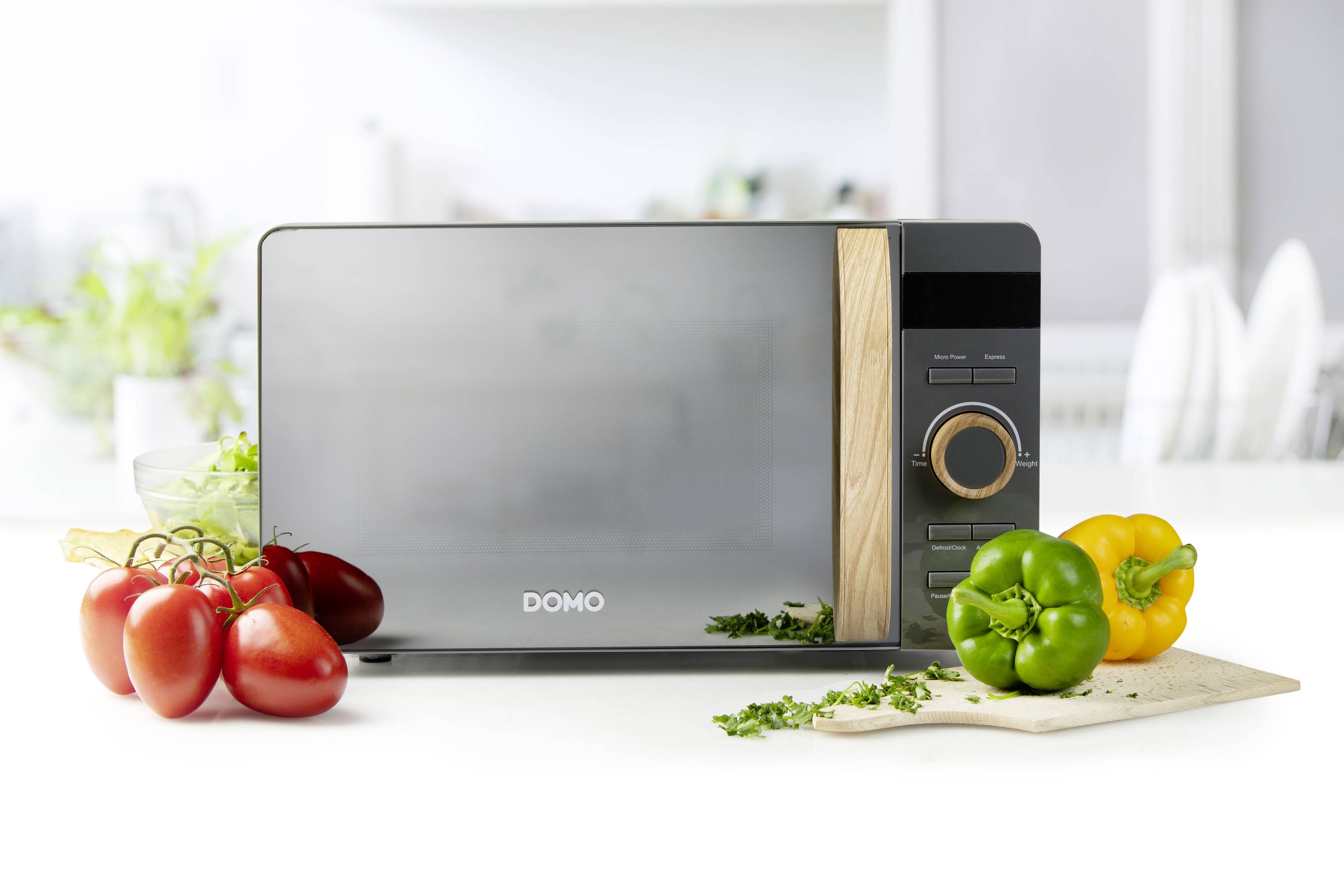 A microwave sits on a kitchen worktop, surrounded by fresh vegetables: tomatoes, peppers and herbs on a wooden chopping board.