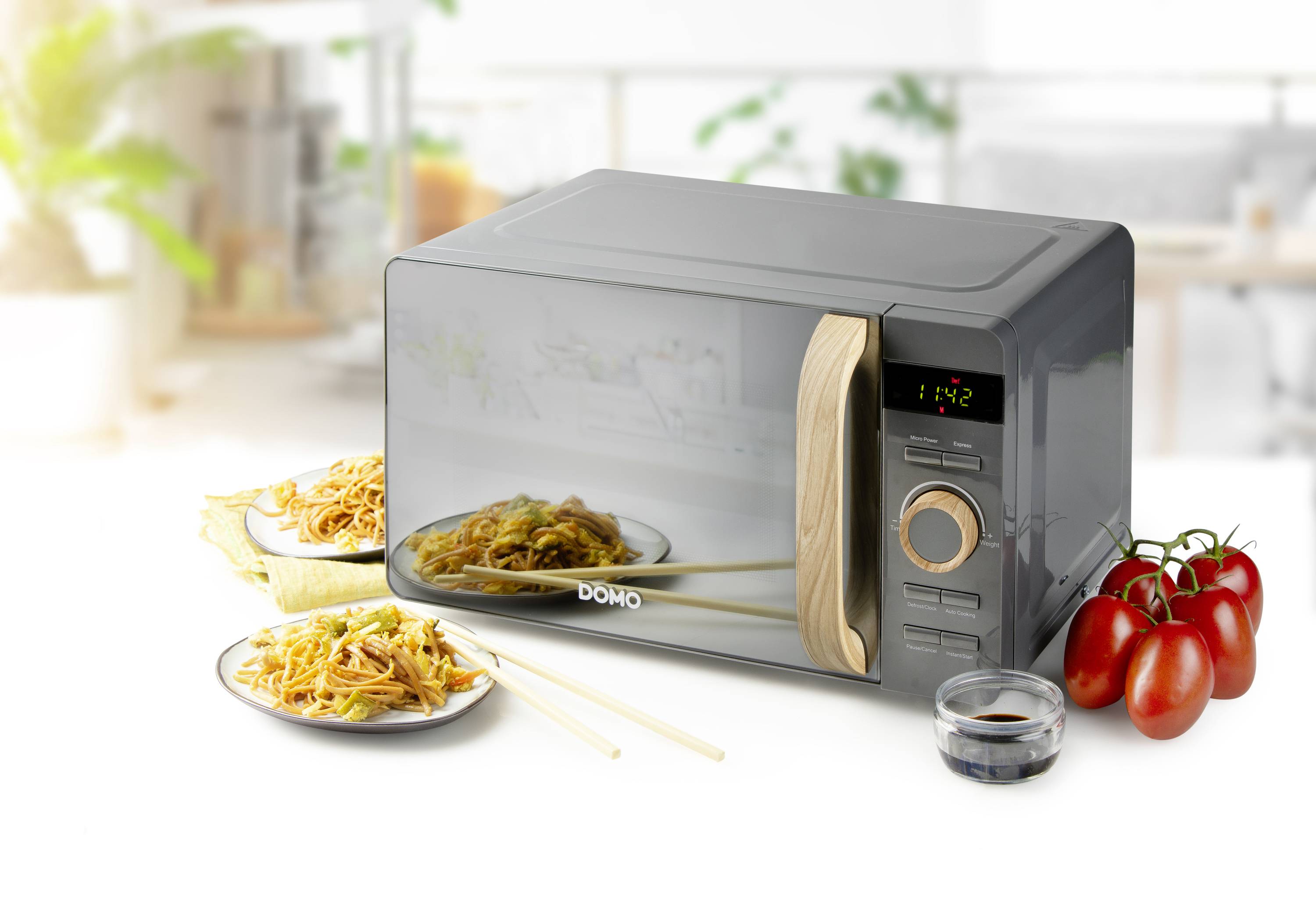 Microwave on a kitchen worktop with an angled wooden handle, with plates of pasta, cherry tomatoes and a bowl of soy sauce beside it.