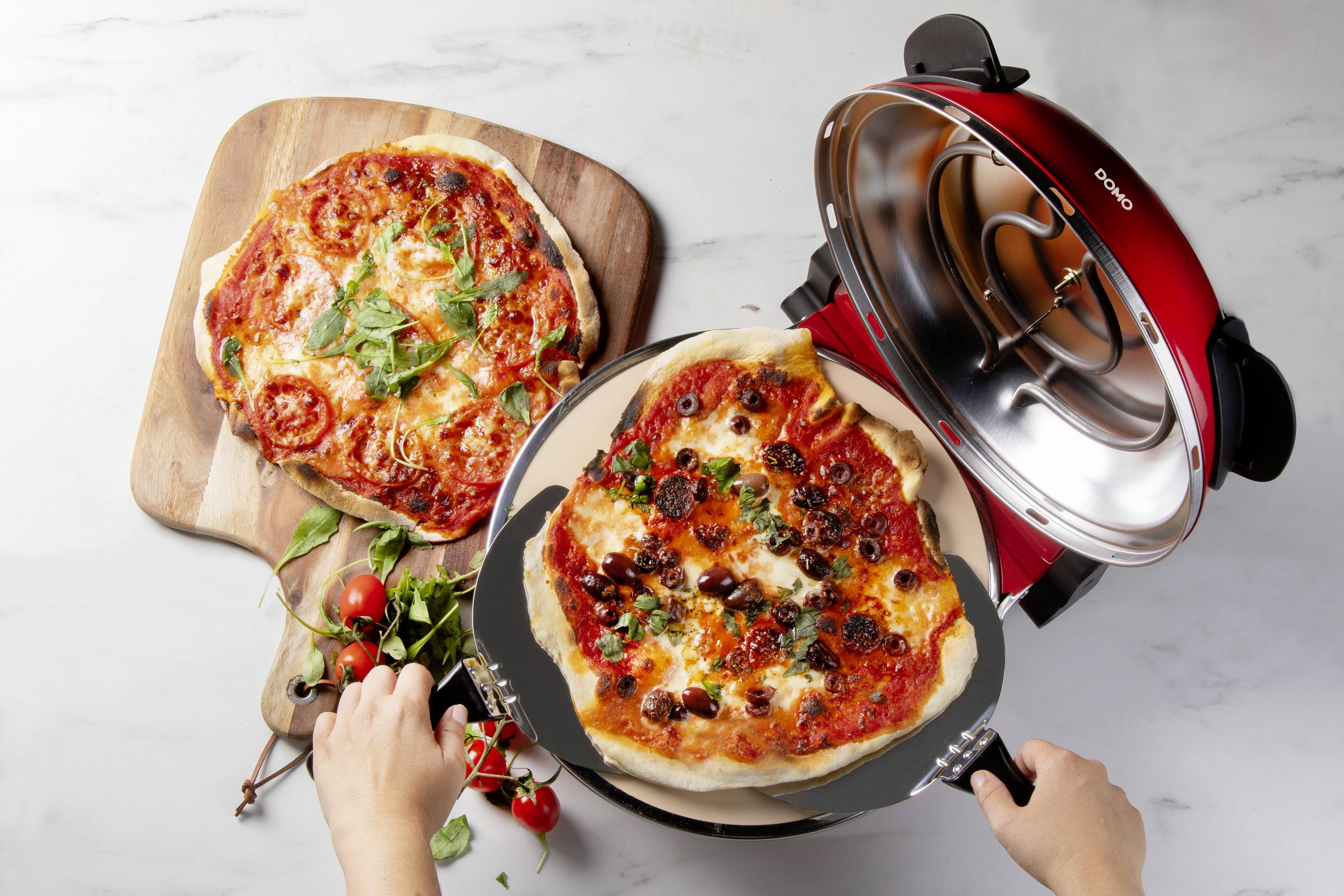 Two pizzas, one freshly baked on a pizza board and the other in an open, red pizza maker. Hands are holding the pizza.