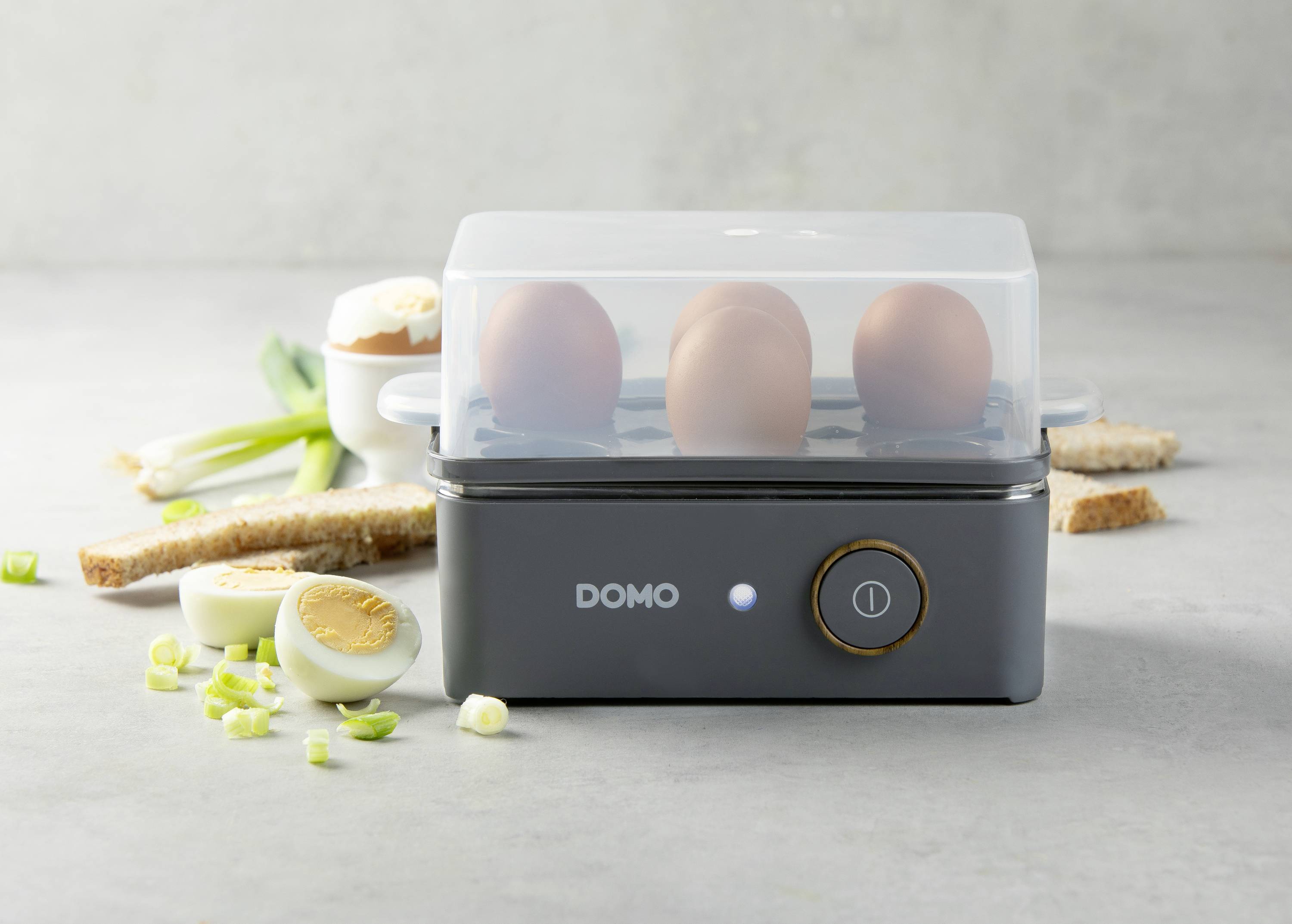 An electric egg boiler with three boiled eggs, a transparent lid, and pieces of toast in the background.