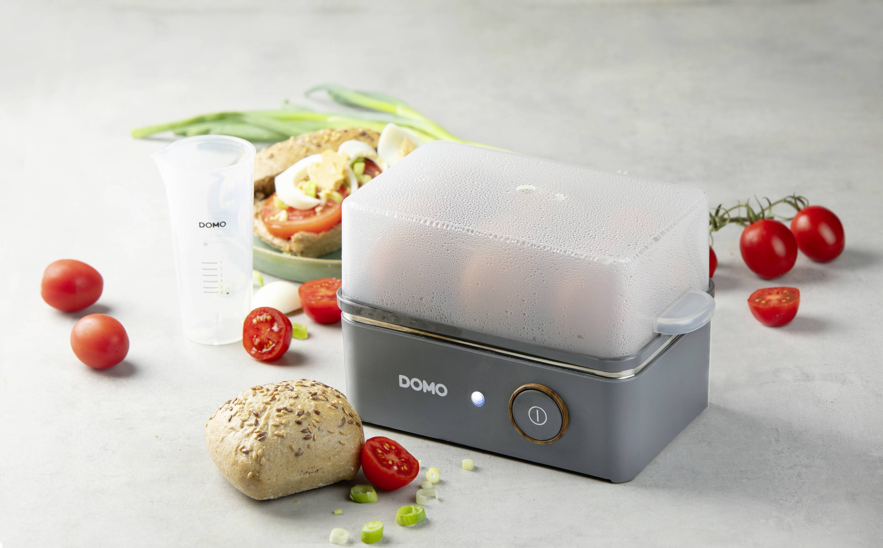 An electric egg cooker with a transparent lid, placed next to tomatoes, a bread roll, and a measuring jug, on a grey table.