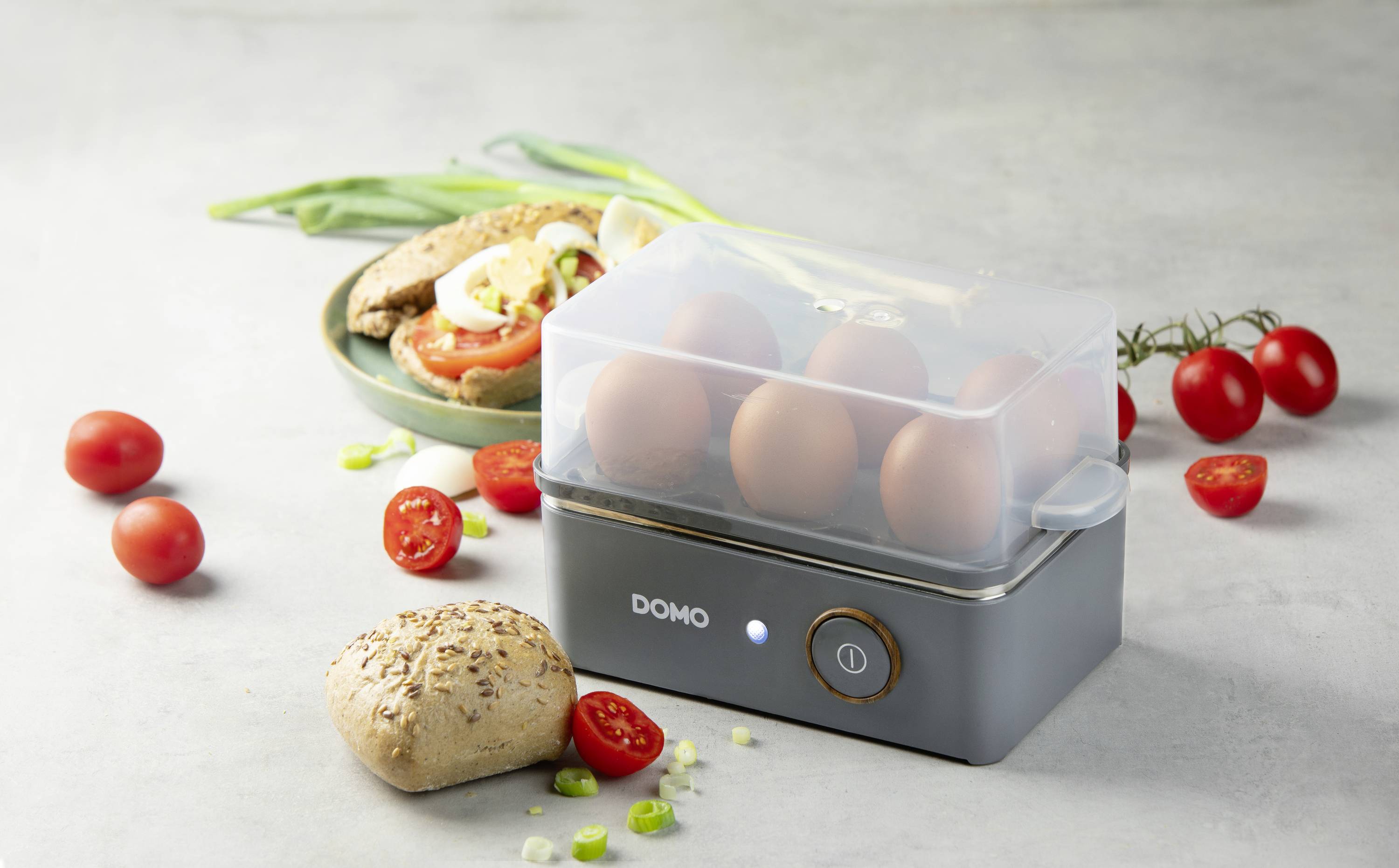 An electric egg cooker with boiled eggs sits on a table. Next to it are cherry tomatoes, a bread roll, and spring onions.