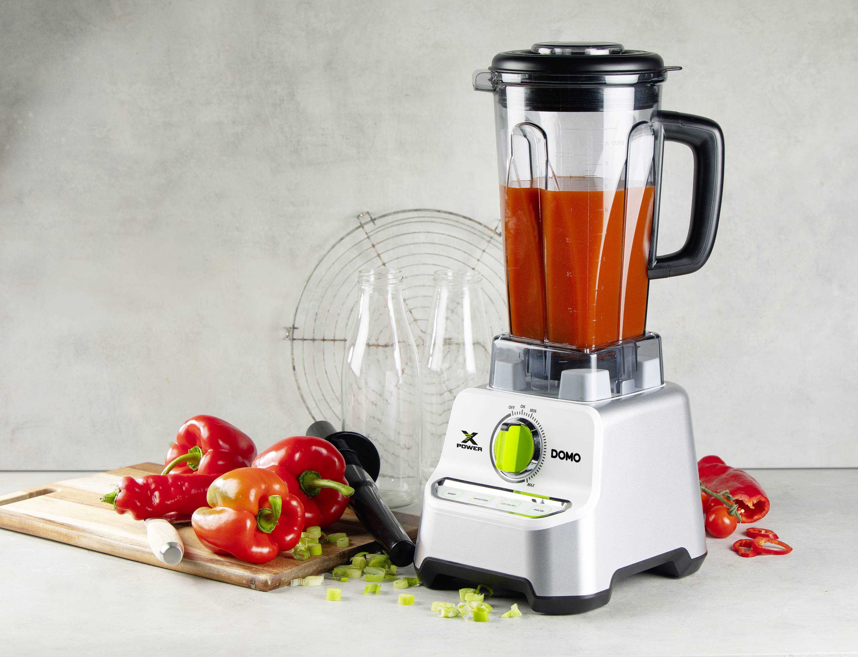 A red blender full of tomato soup stands next to chopped peppers and spring onions on a wooden board in a kitchen.