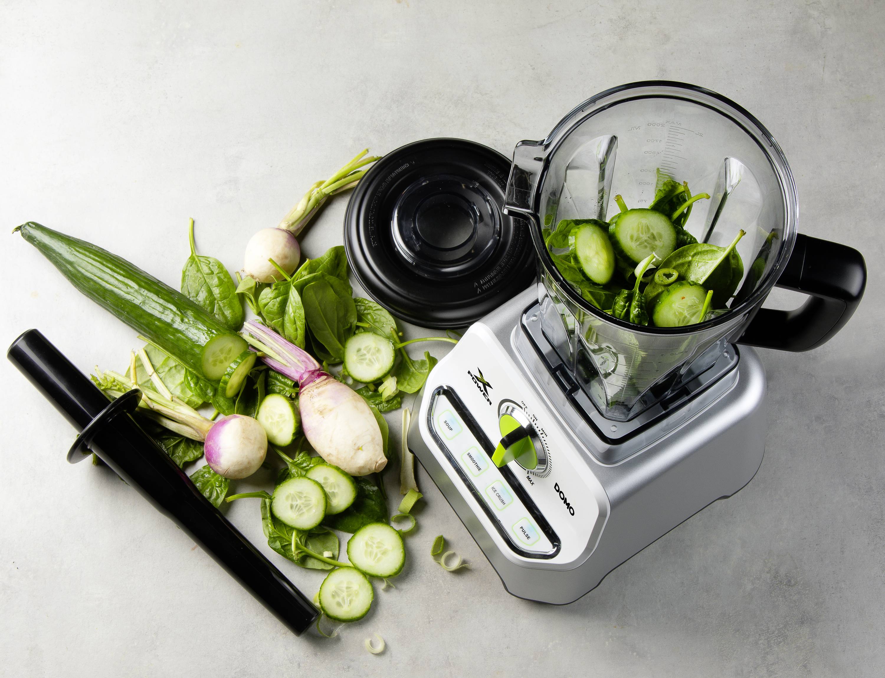 Blender with an open container, filled with chopped cucumbers. Beside it, fresh vegetables, including cucumbers, radishes, and spinach.