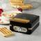A black waffle maker by Domo on a kitchen worktop, beside a bowl of fruit and freshly baked waffles in the background.
