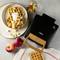 Waffle iron with a waffle on a wooden table, plus a plate with two waffles, apple pieces and cream. Two whole apples and extra apple pieces in a bowl.
