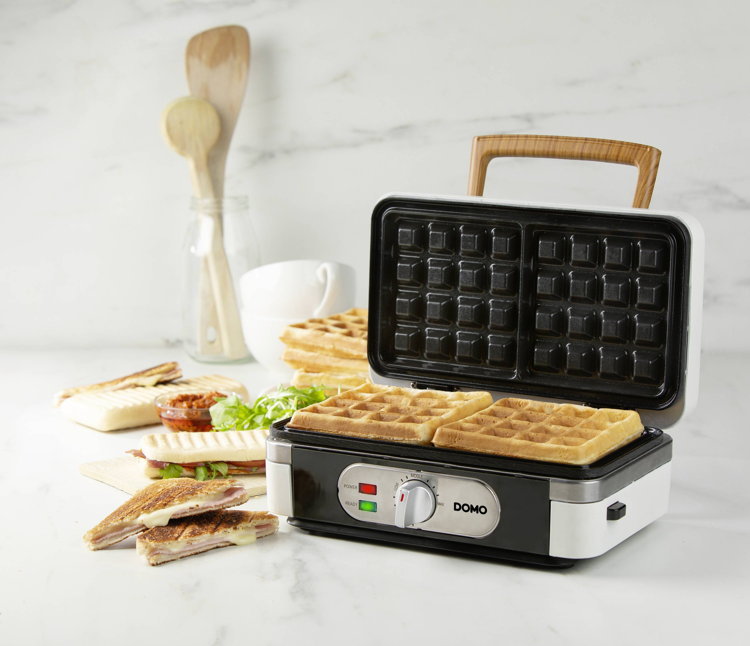 An open waffle maker with two freshly made waffles inside. Beside it are sandwiches, a wooden spoon set and a cup against a marble background.