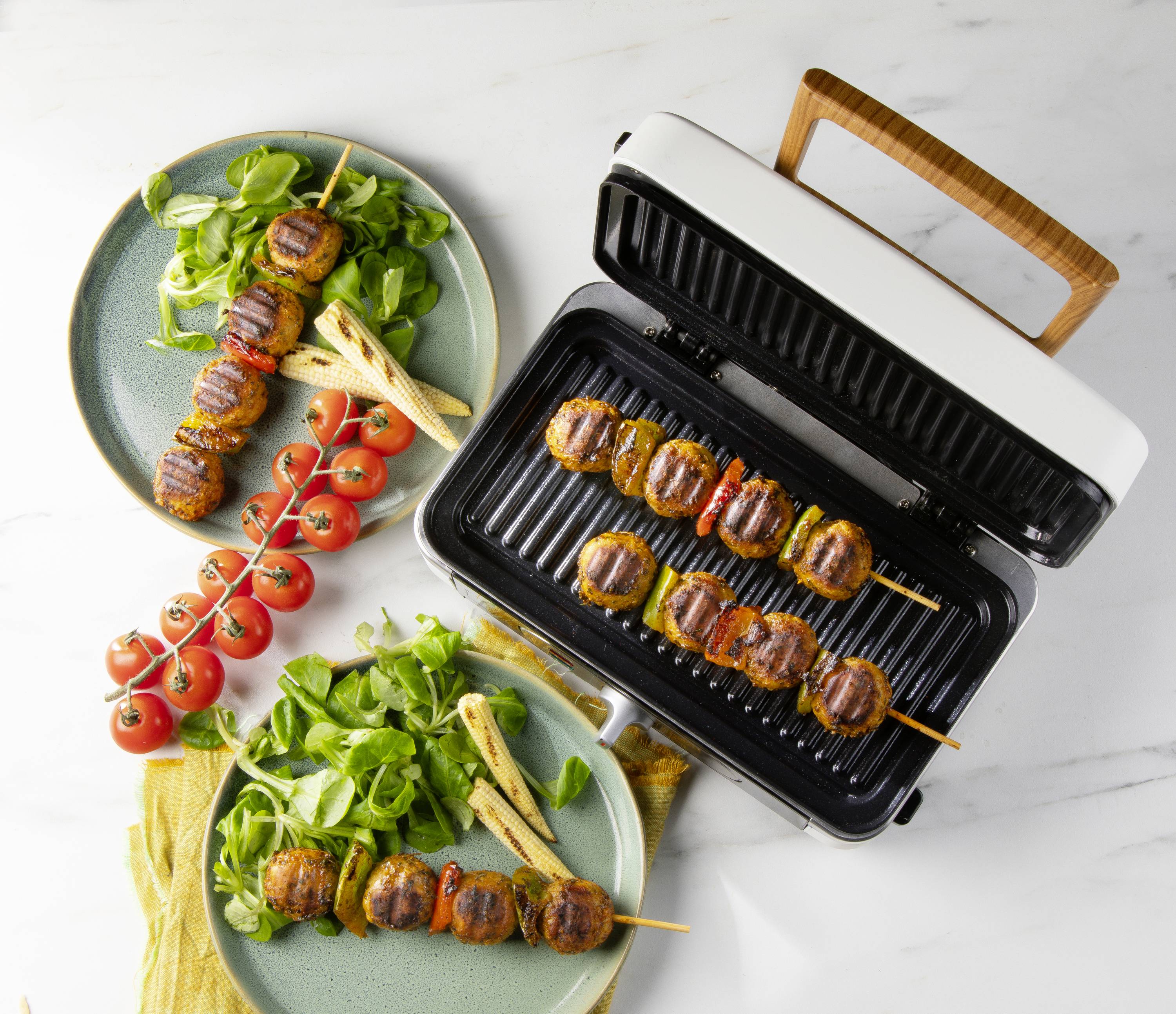 Two plates with grilled vegetable skewers and salad beside an electric barbecue with additional skewers on a marble table.