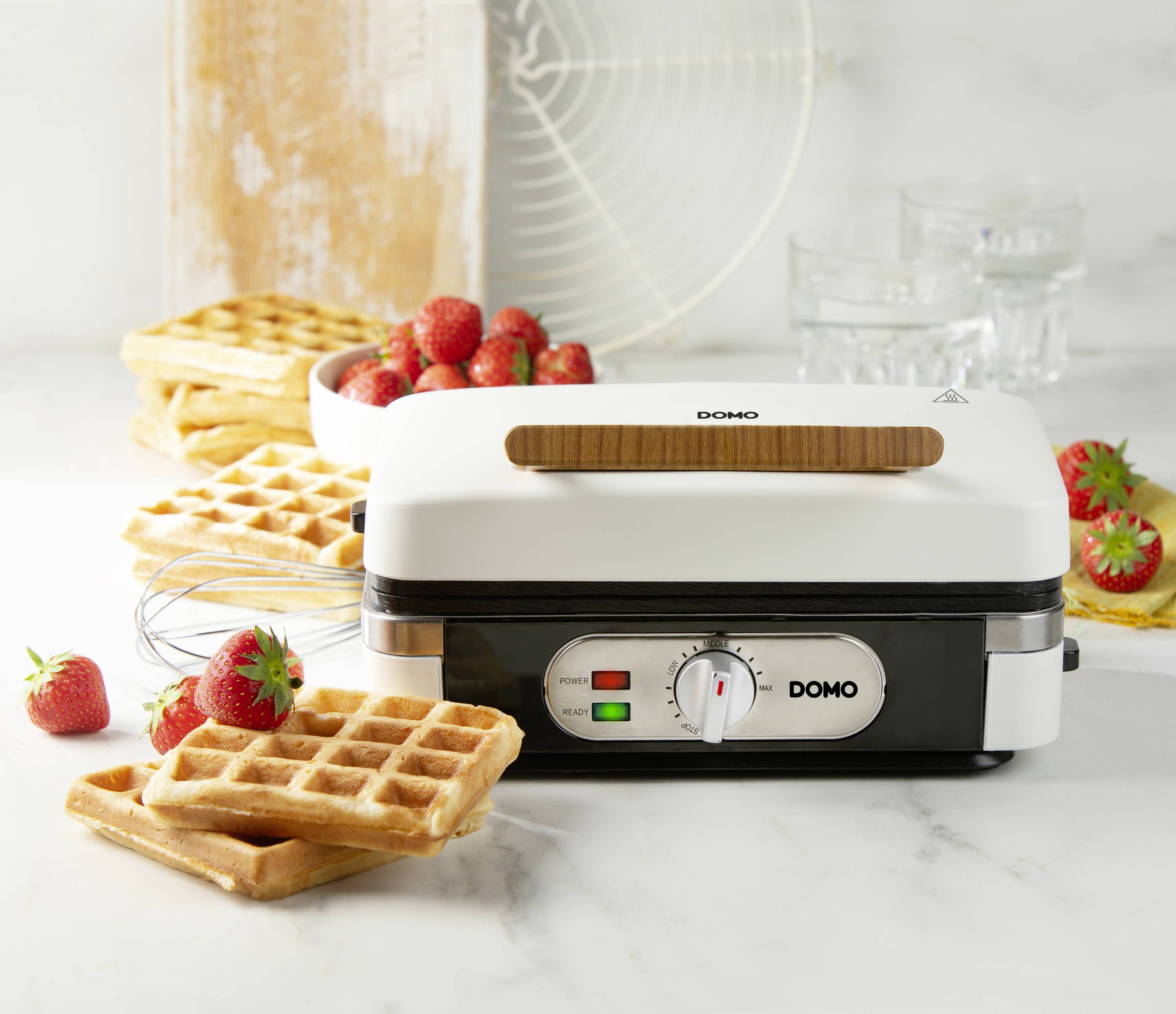 A waffle iron on a kitchen worktop, surrounded by freshly baked waffles, ripe strawberries, and a whisk.