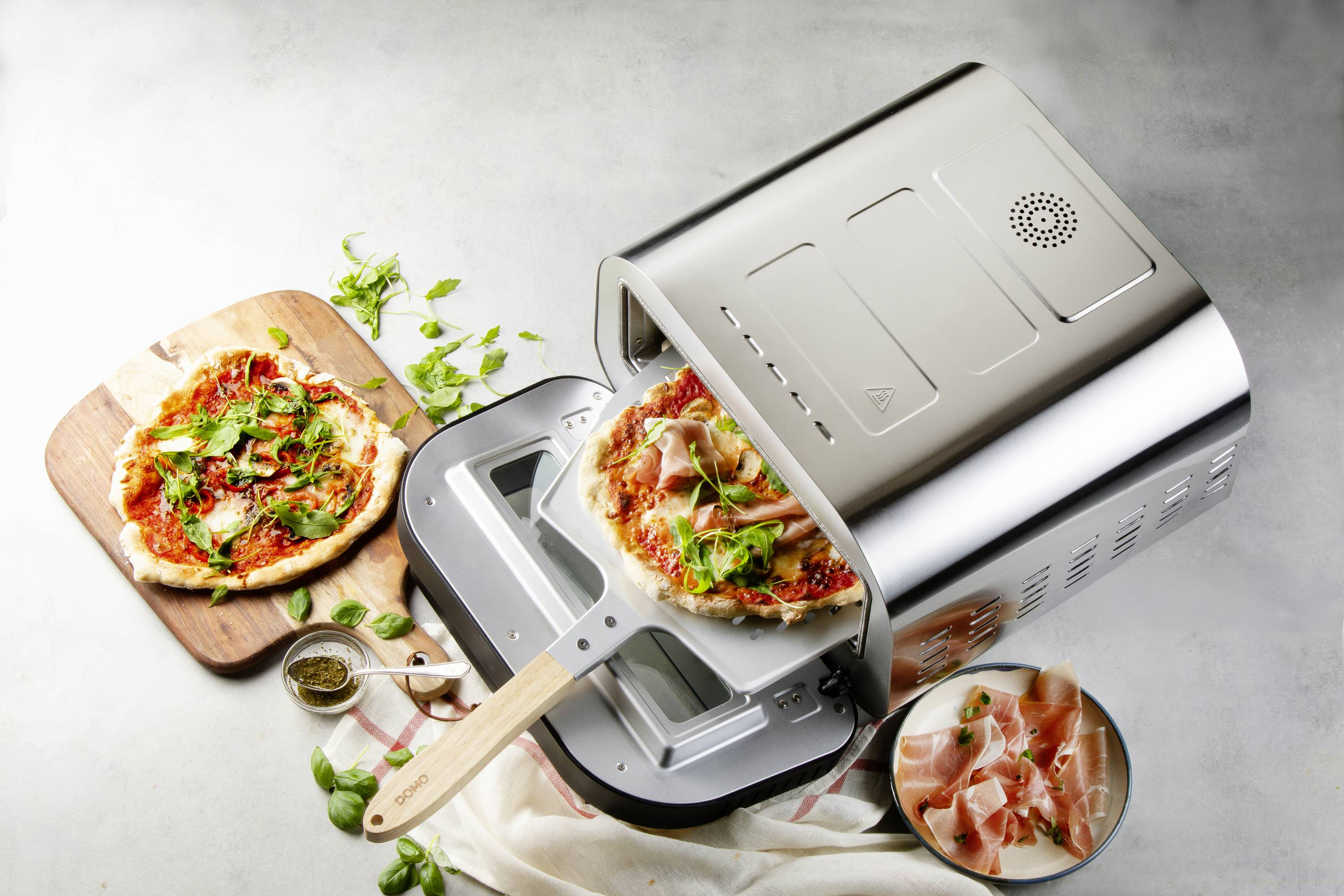 A pizza oven on a worktop is baking a pizza with ham and basil. Next to it, a finished pizza lies on a wooden board.