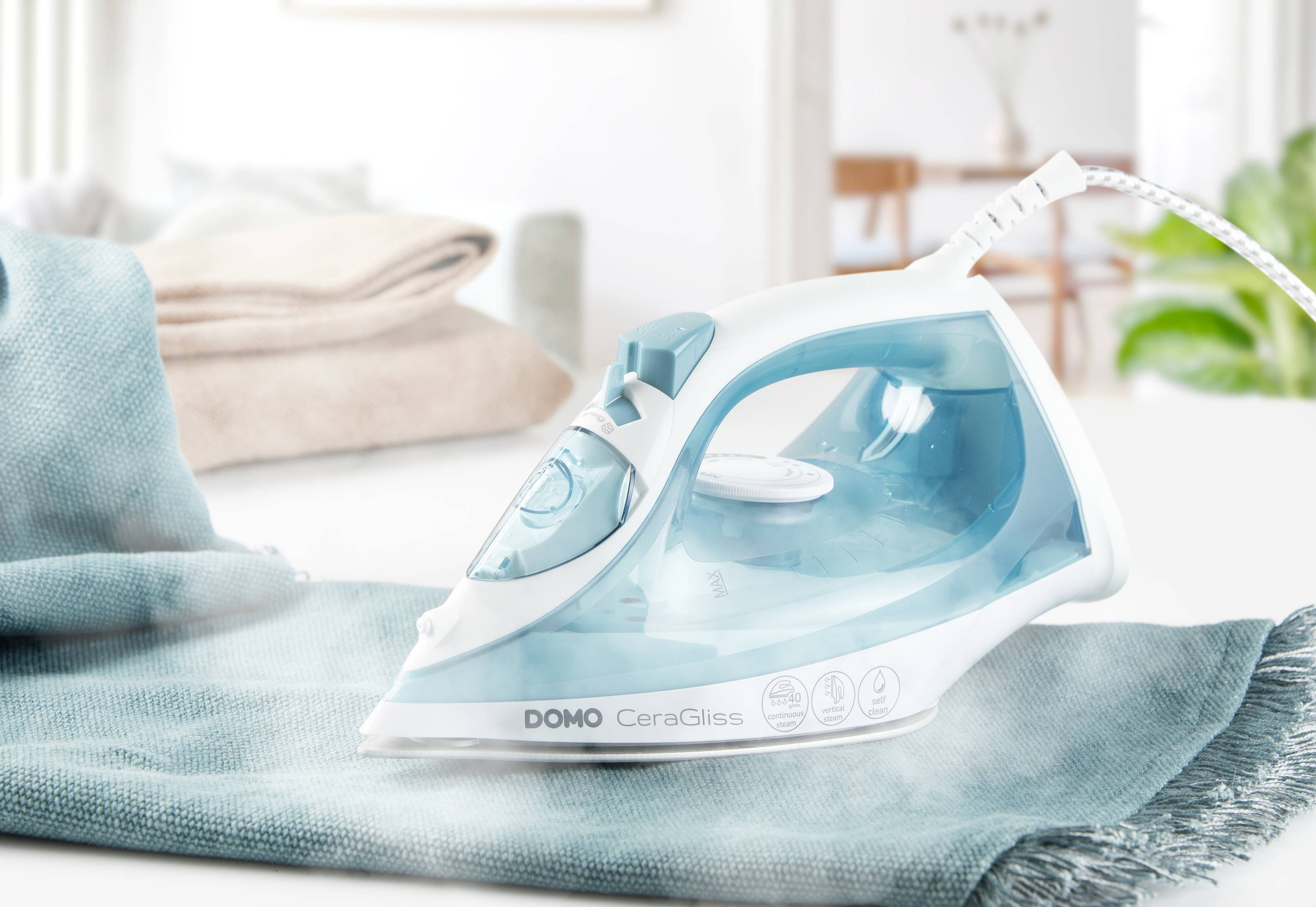 A steam iron on an ironing board is pressing a blue cloth. Blurred folded towels can be seen in the background.