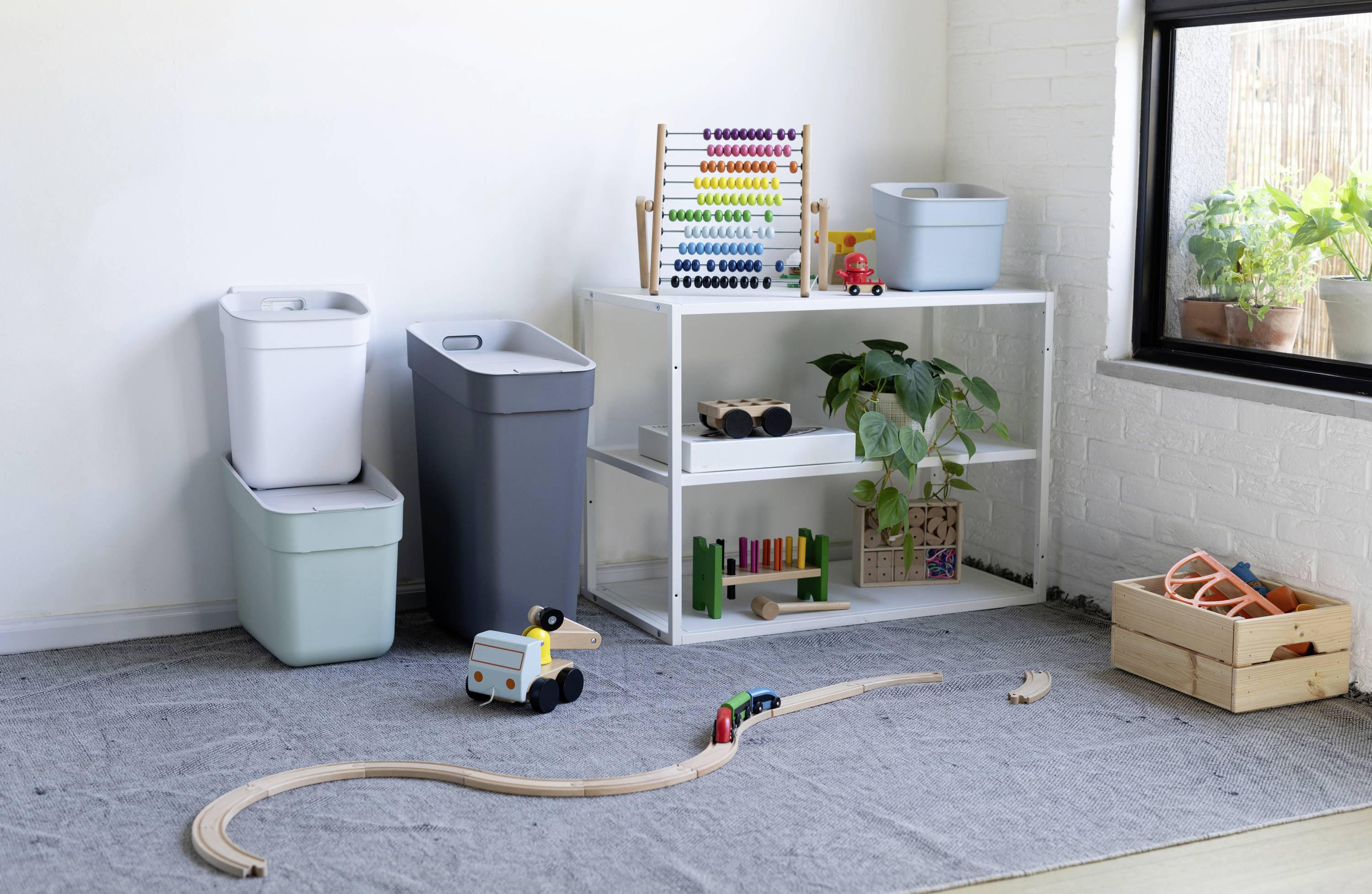 A playroom with a toy shelf, a wooden train set on the floor, and plants on a windowsill in a bright room.