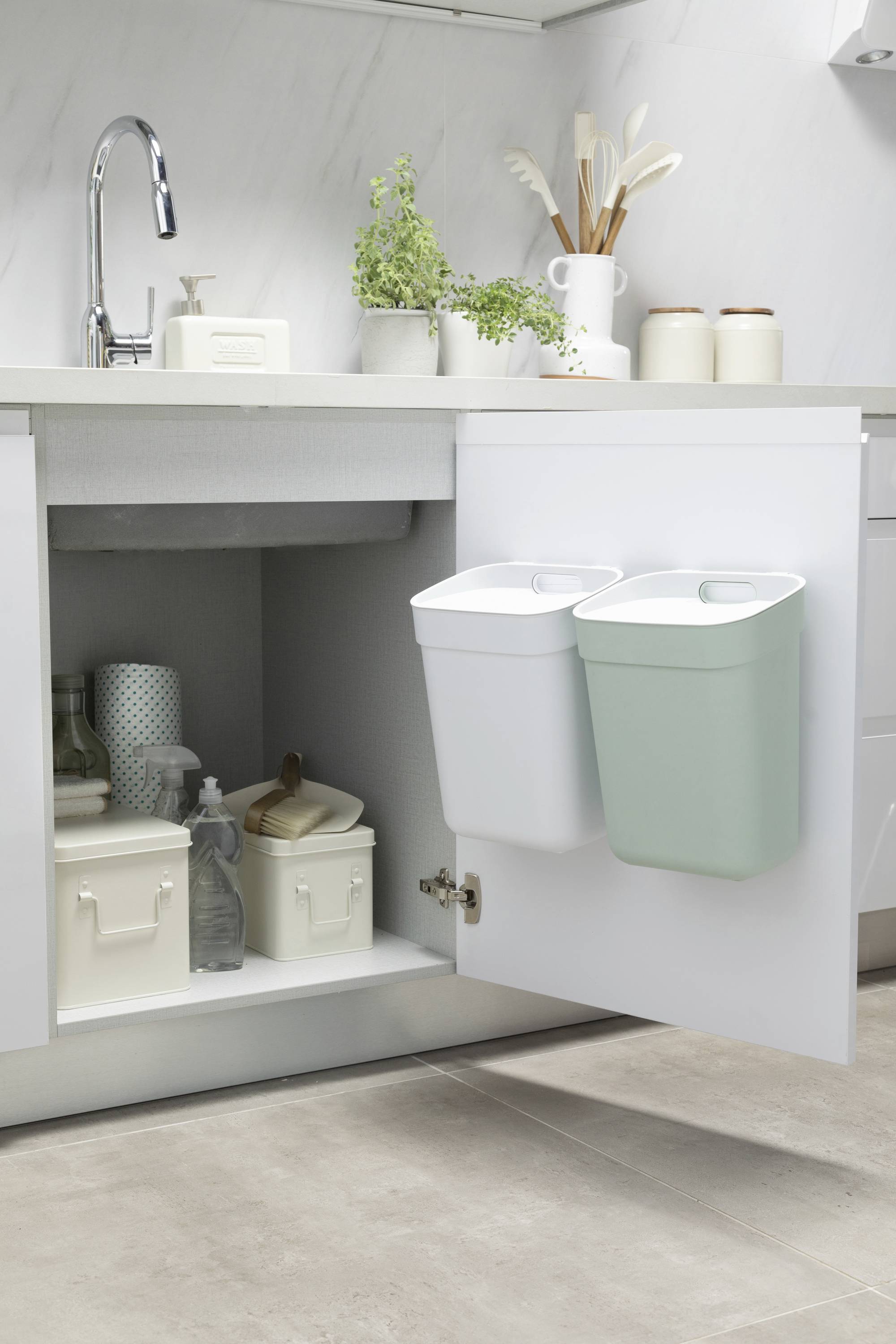 Kitchen cupboard with two bins on the inside of the door. Additional storage containers underneath. Plants beside the sink at the top.