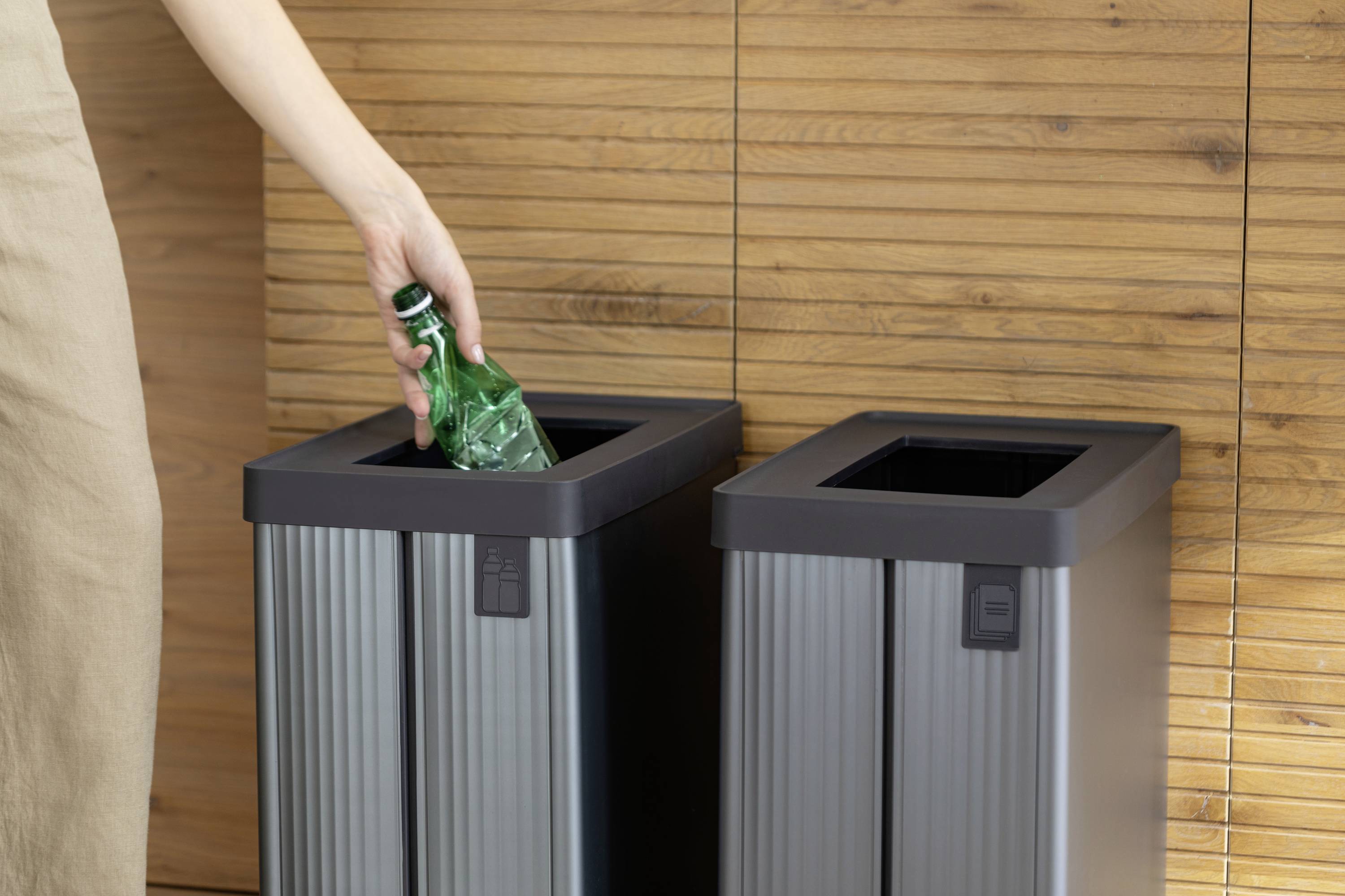 A person is throwing a green glass bottle into a waste bin with a segregation system. The background consists of wooden wall panelling.