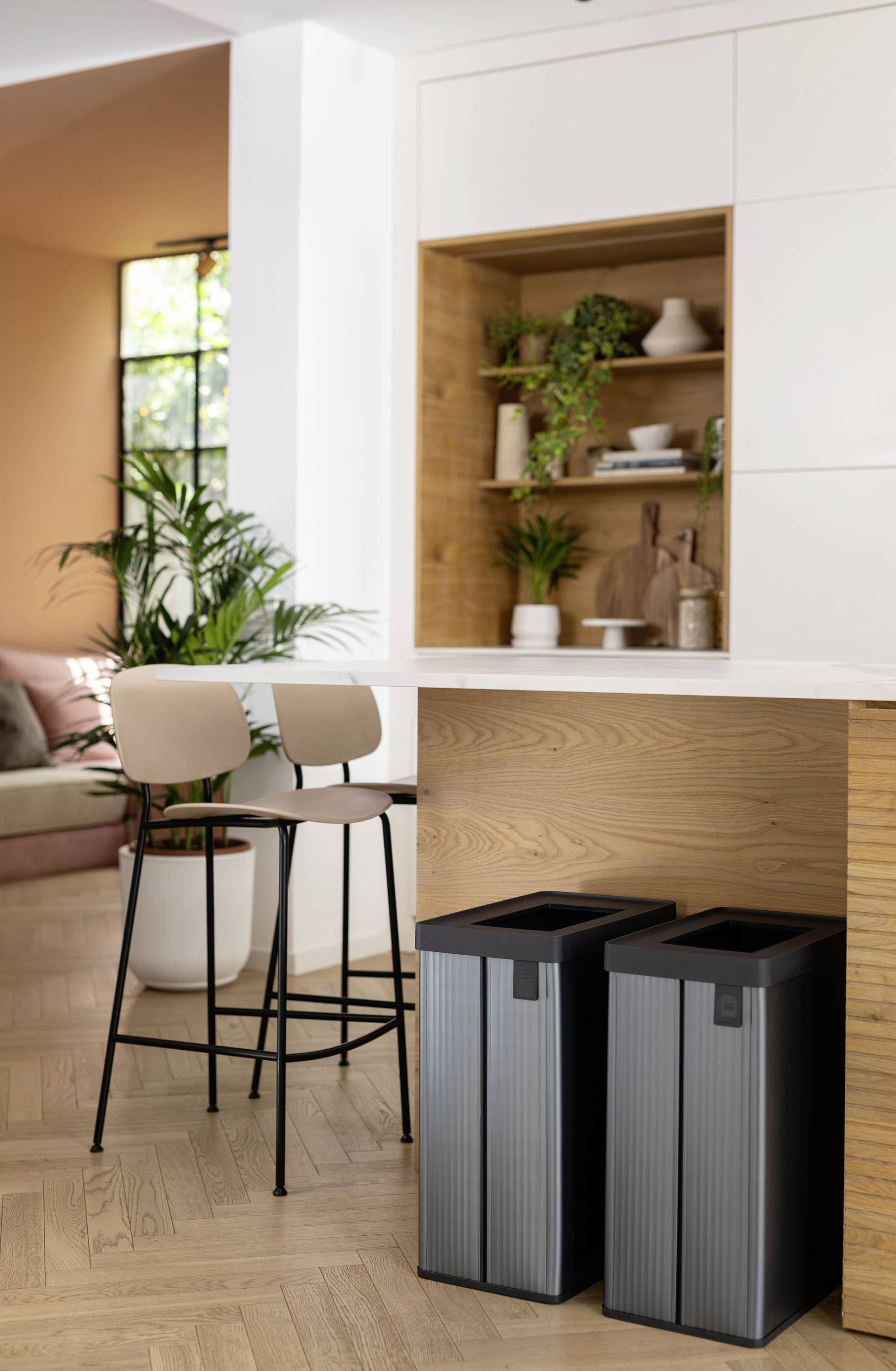 Modern kitchen area with two high stools at a breakfast bar and two rectangular bins underneath; plants and wooden shelves in the background.