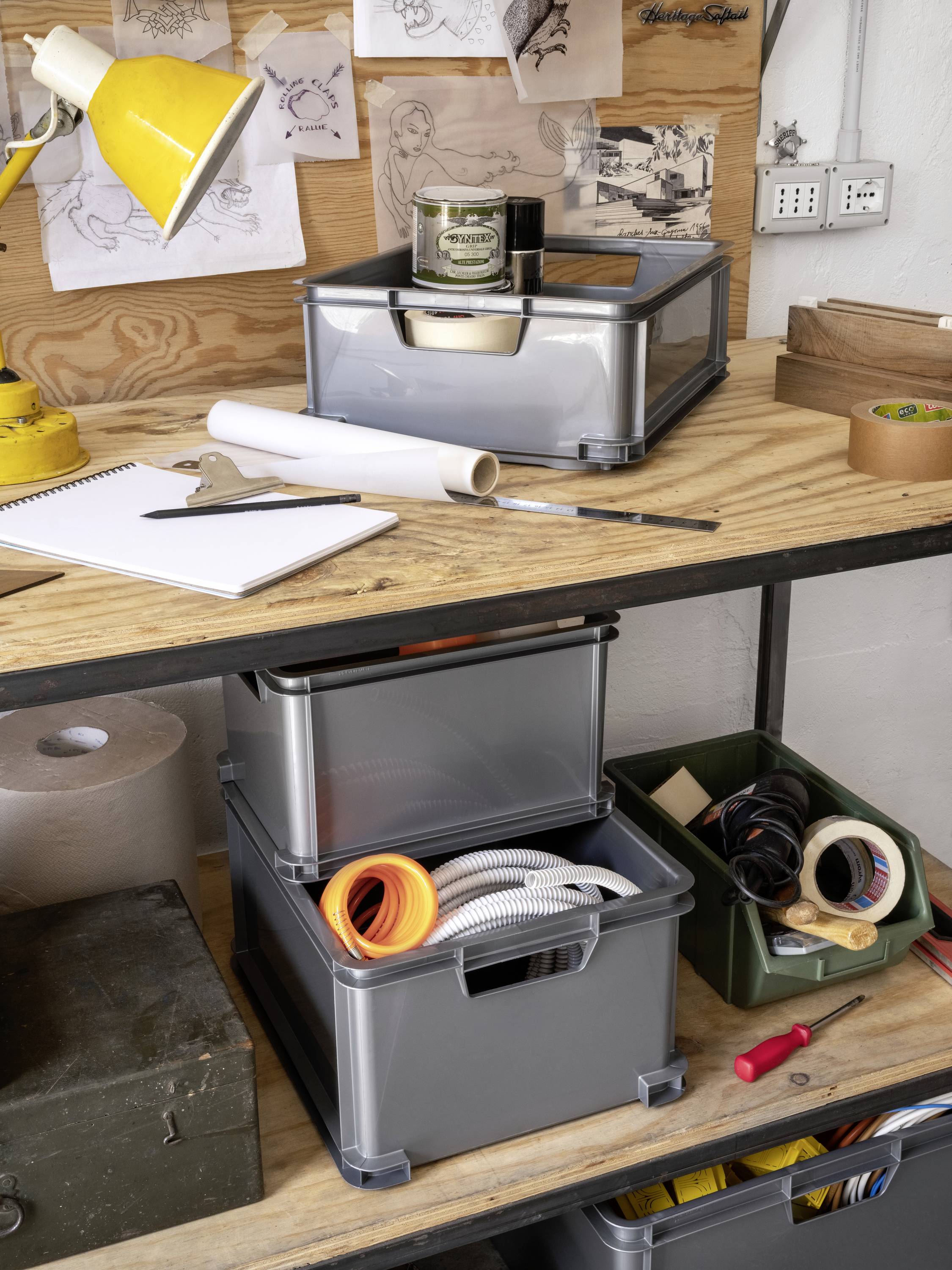 A workshop table with storage boxes, notebook, lamp, and tools. Drawings and notes are attached to the wall.