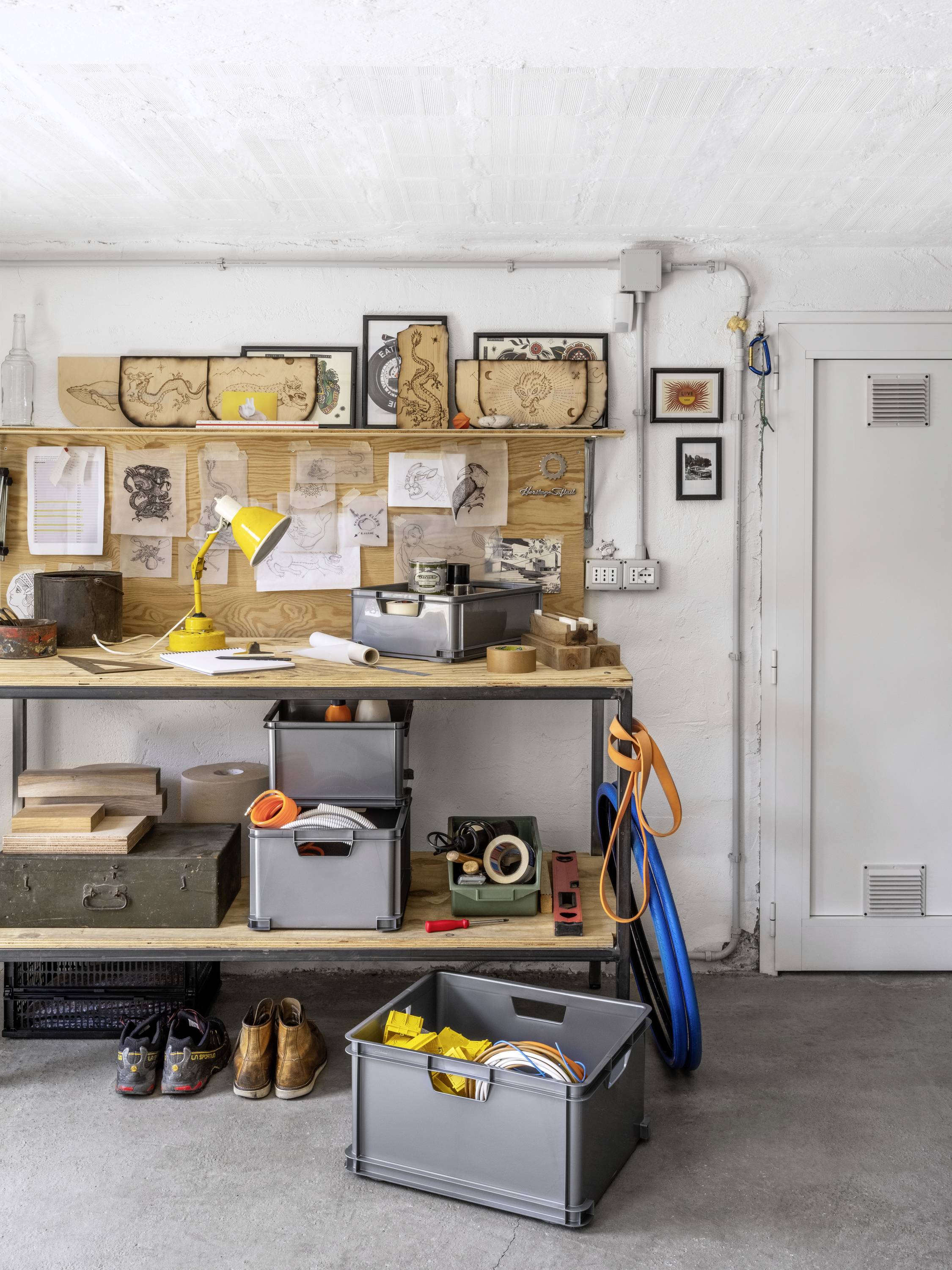 Workshop with a wooden workbench, featuring a yellow lampshade, tools, and drawings on the wall; plastic boxes below with cables and tools.