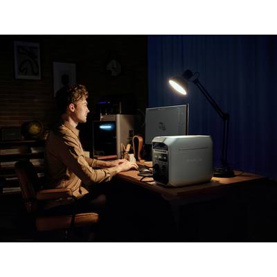 A man sits in a dark room at a desk, with a computer and a generator on it, illuminated by a lamp.