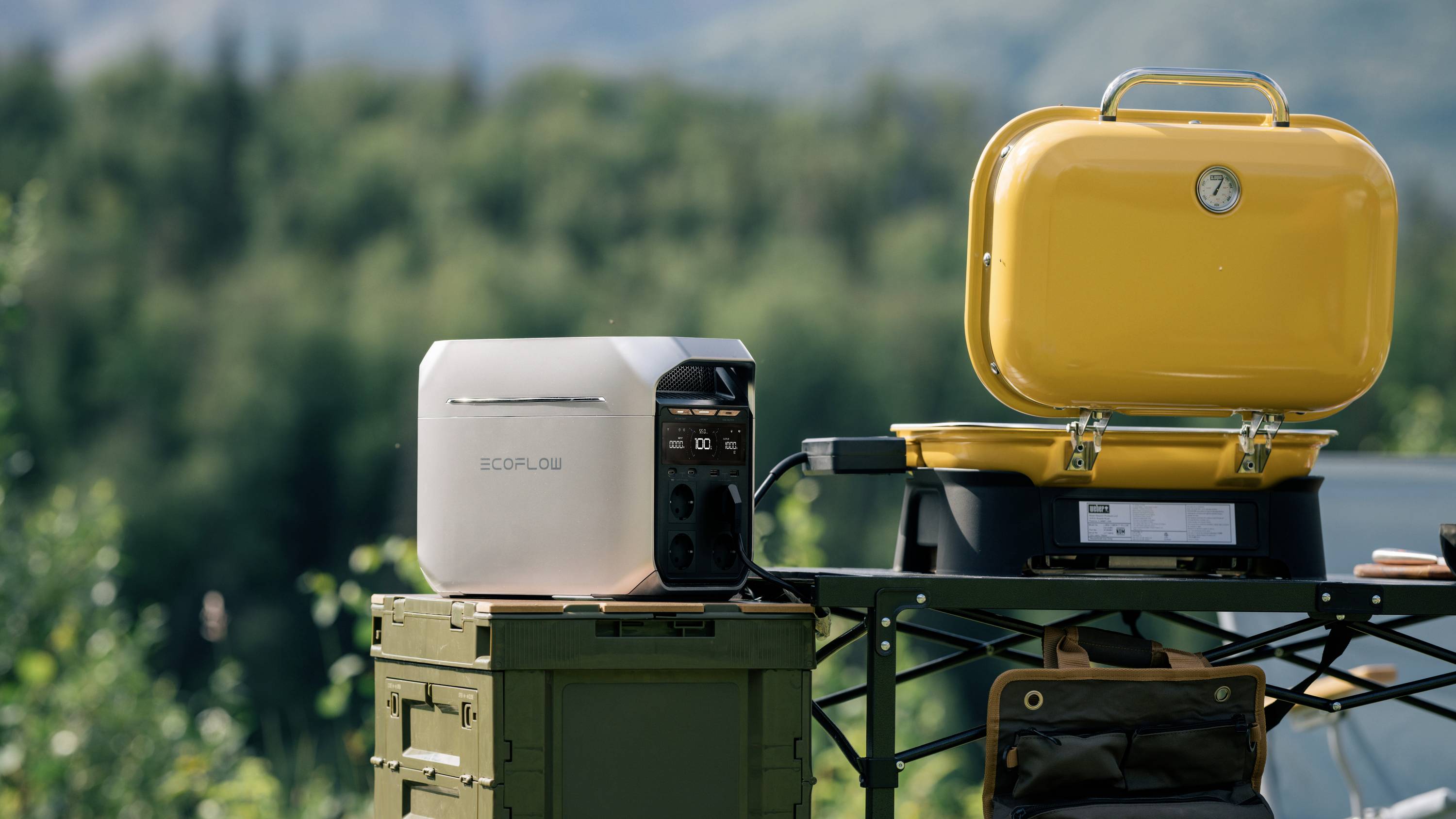 A portable EcoFlow power station sitting on a table next to a yellow barbecue grill outdoors. Trees and mountains are visible in the background.